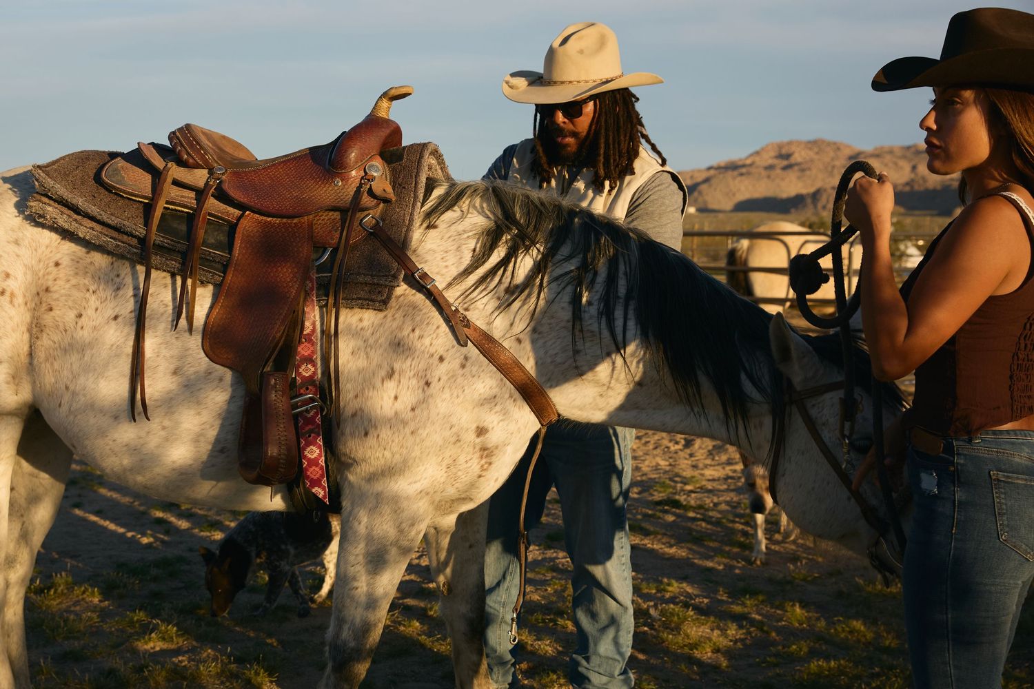 Two cowboys in hats saddle a gray horse in an open desert landscape at golden hour.