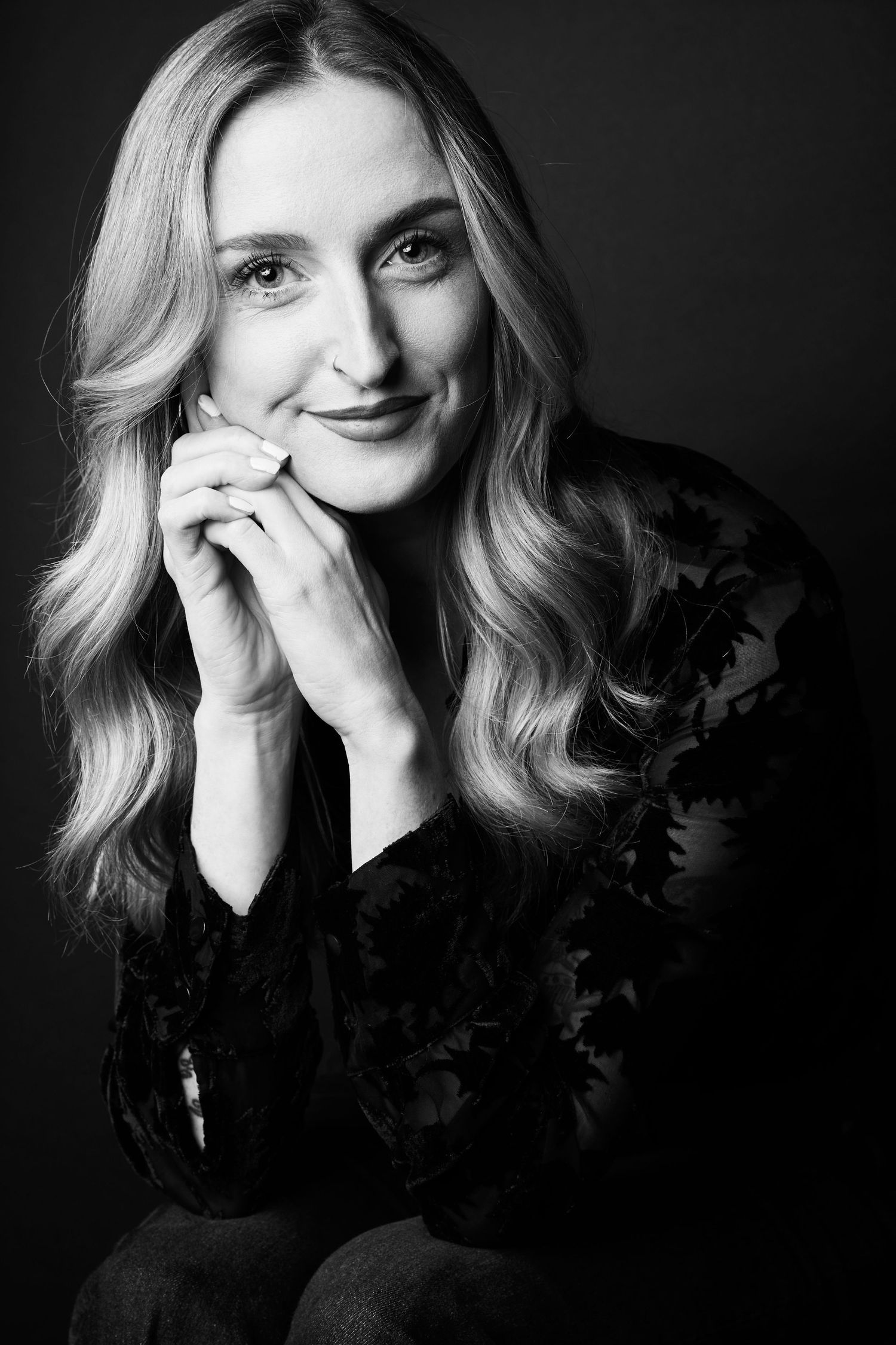 Black and white portrait of a smiling blonde woman in floral top, hands clasped under chin.