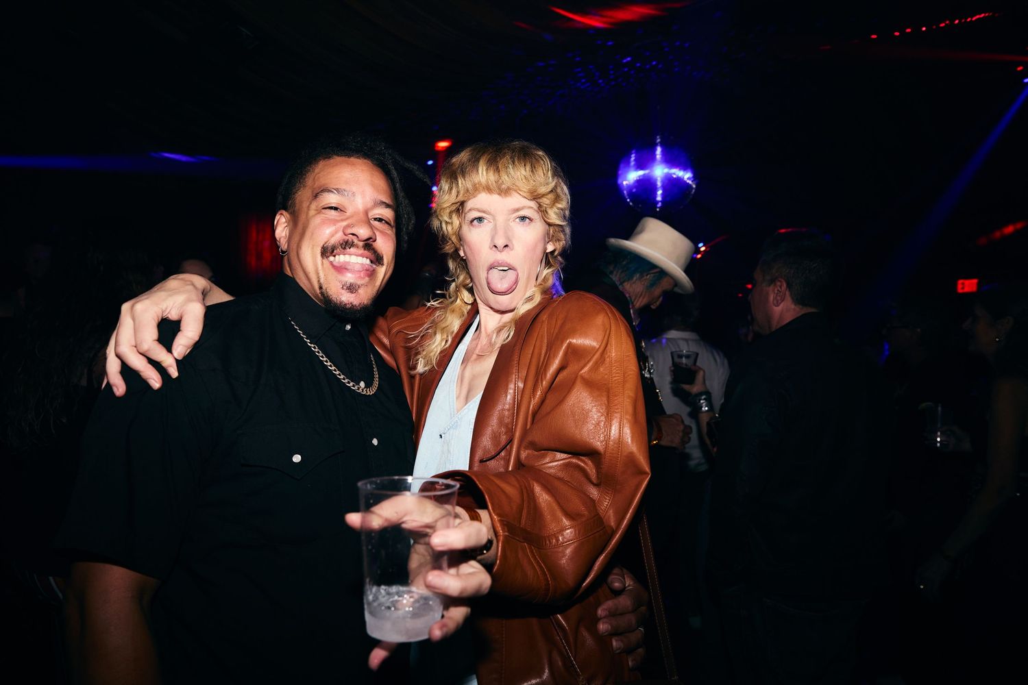 Two people smile and pose together at a nightclub with one wearing a brown leather jacket and another in dark clothing.