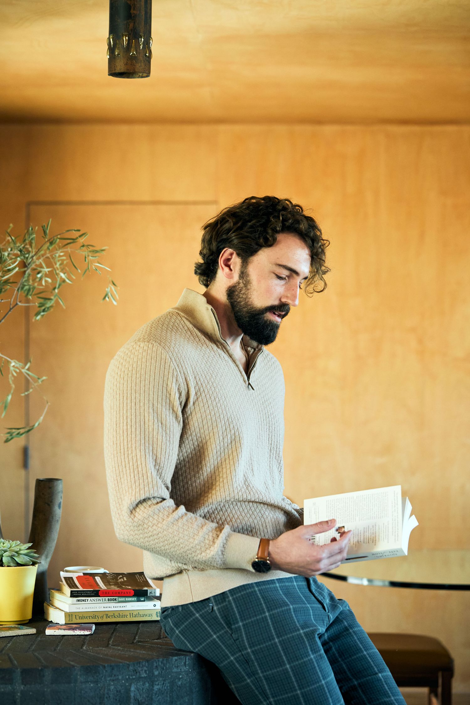 Person in a beige sweater sitting and reading a book in a warm lit room with houseplant and wooden walls.