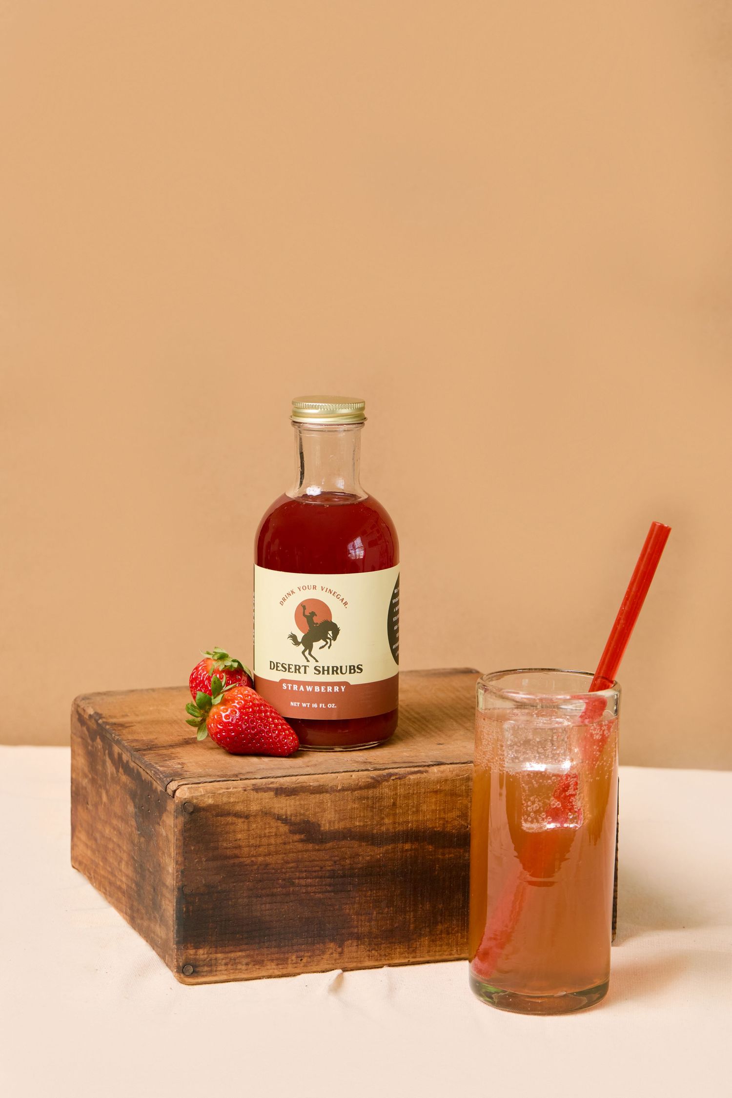 Dumey Shrubs strawberry syrup bottle beside a fresh strawberry and a tall glass of pink cocktail with red straw.