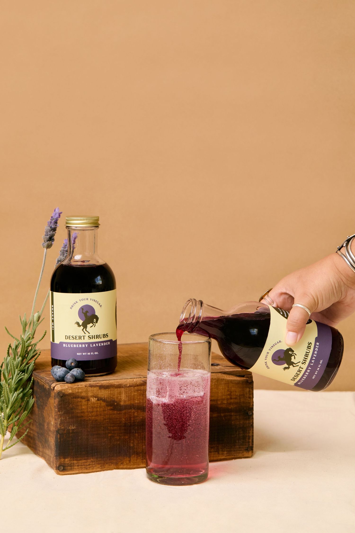 Hand pouring Desert Shrub blueberry lavender syrup into an iced glass, with bottle and fresh herbs on wooden blocks.