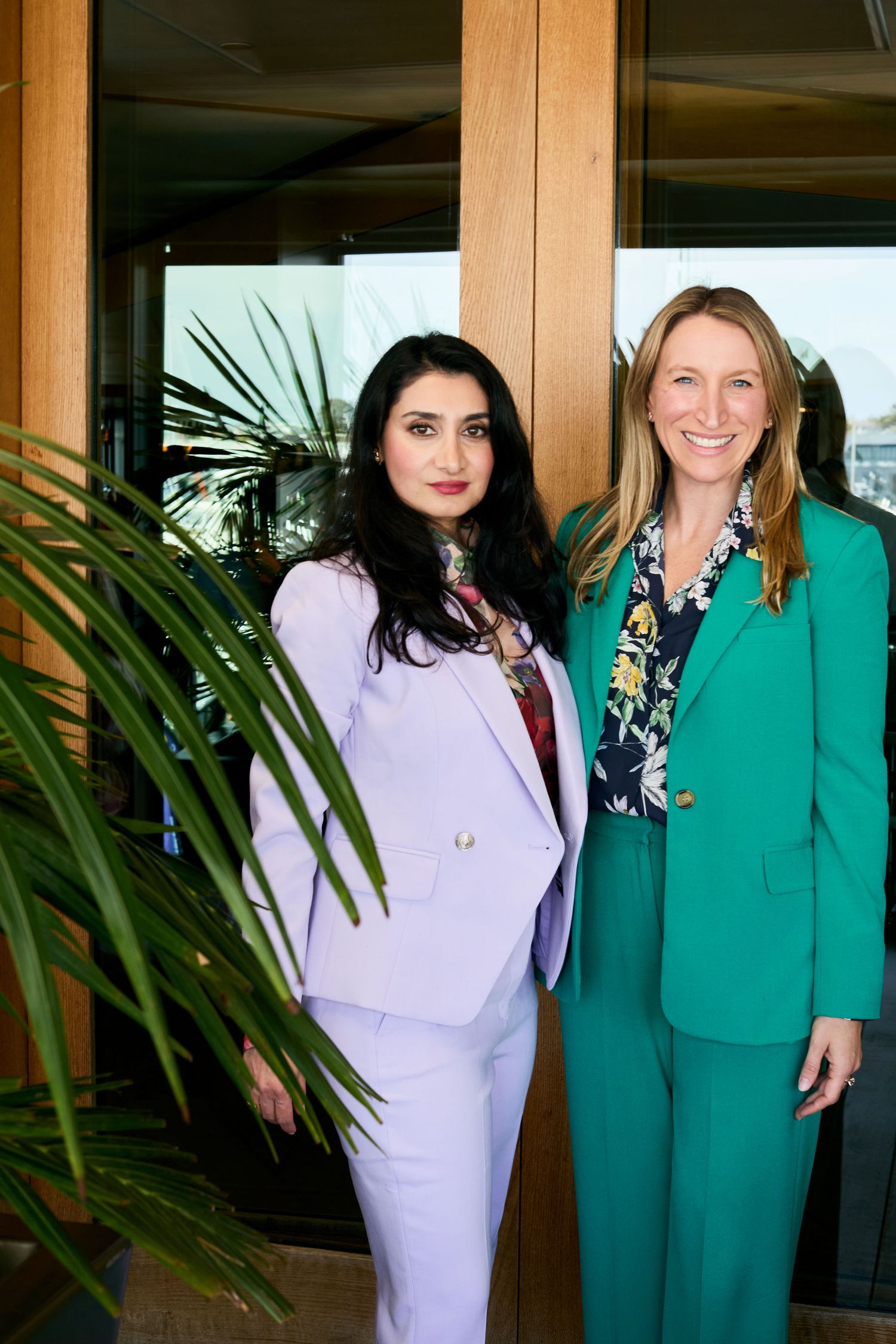 Two professionals in stylish business suits pose together in front of tropical plants in an indoor setting.