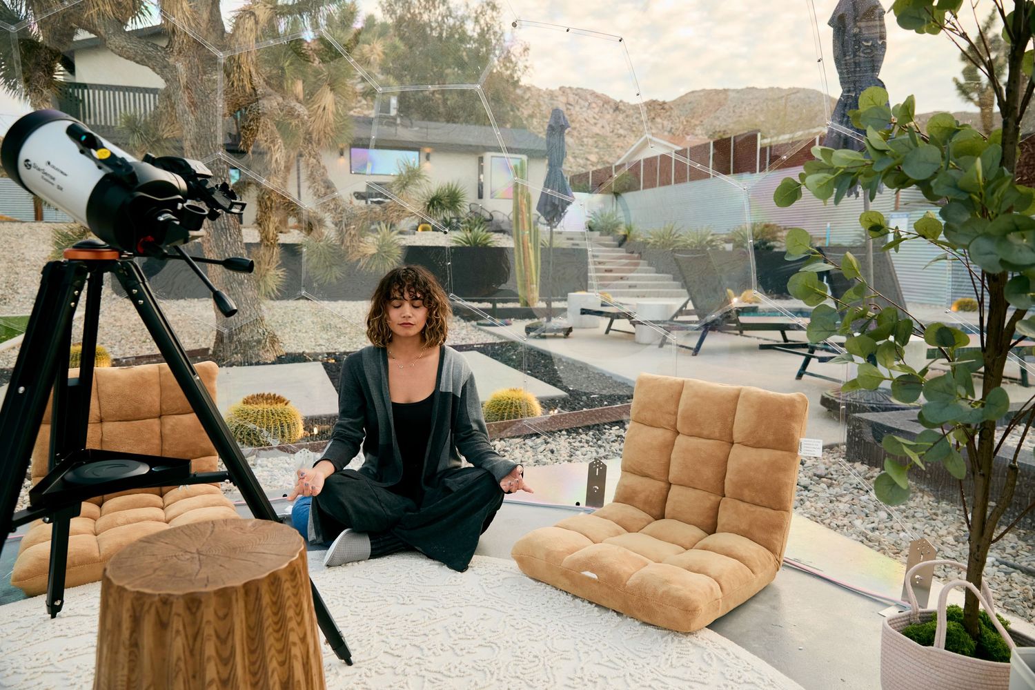Woman meditating in lotus pose outdoors beside a telescope and cozy lounge chair in a desert patio setting.