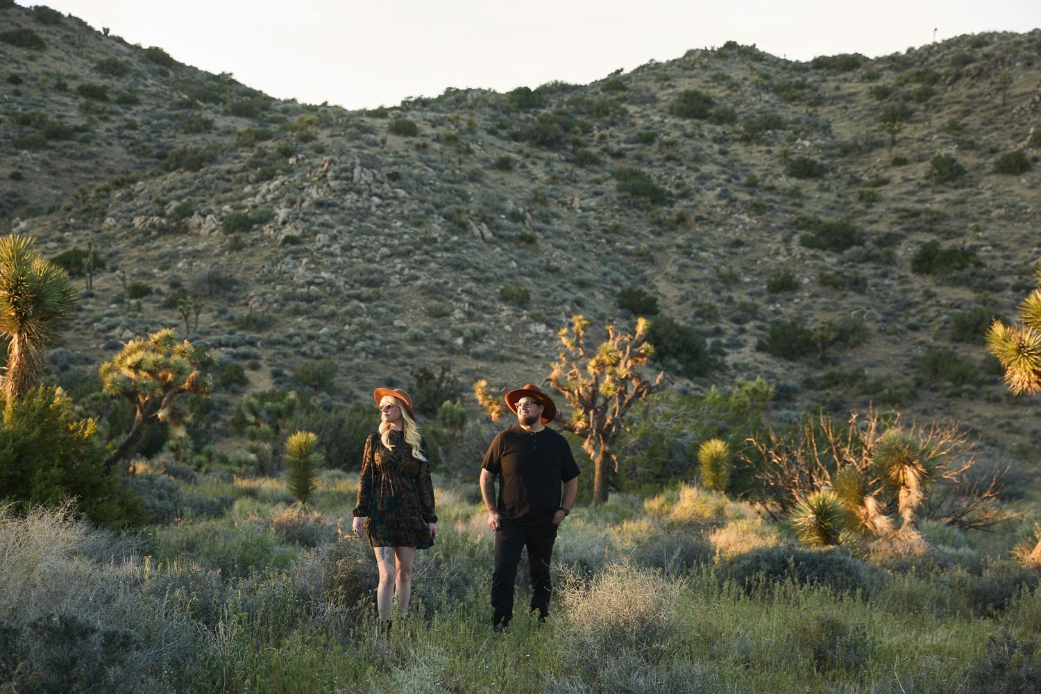 Two people standing in a desert landscape with Joshua trees and scrub brush against a rocky hillside at dusk.