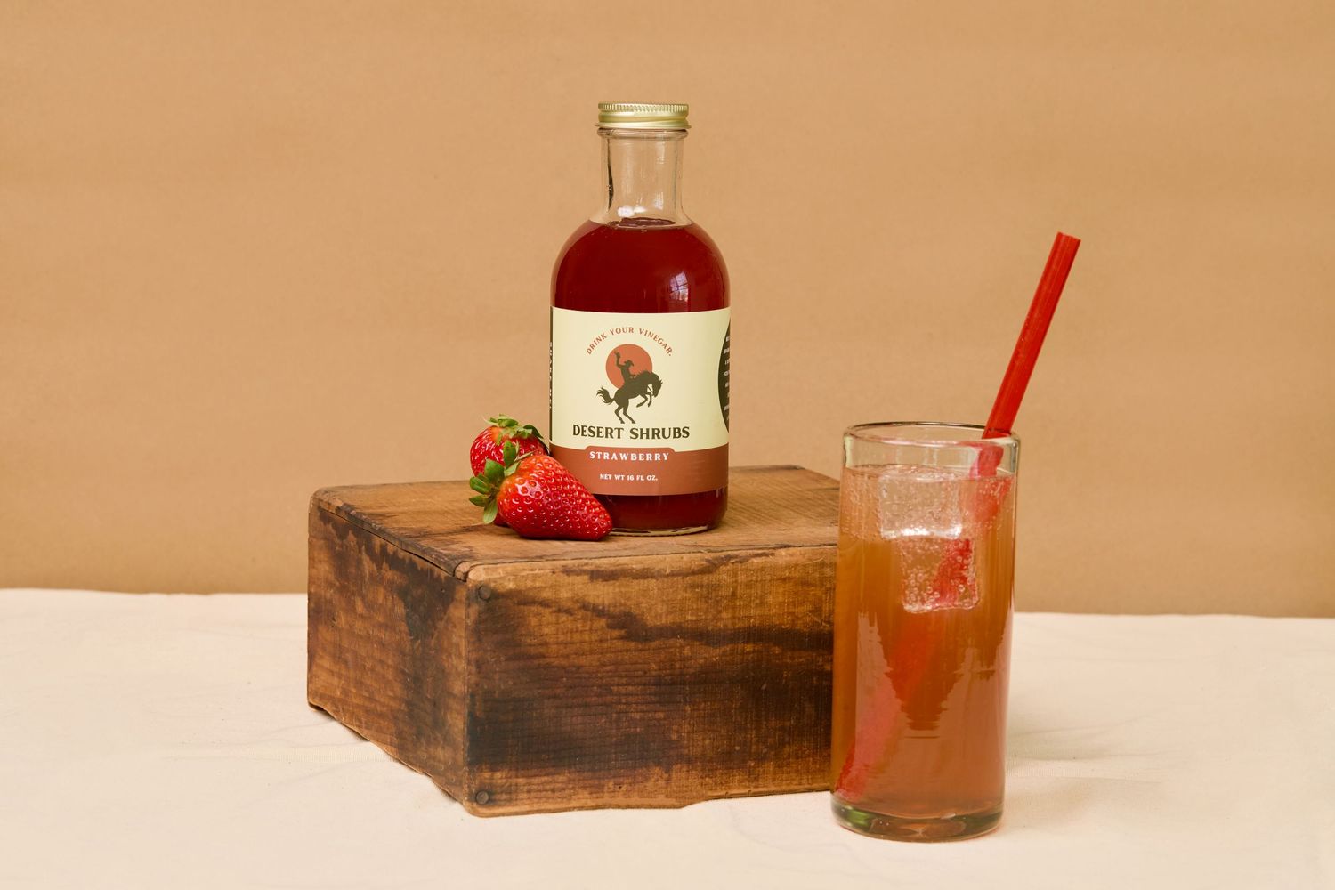 Strawberry simple syrup bottle beside a fresh strawberry and a tall glass of iced strawberry drink with red straw.