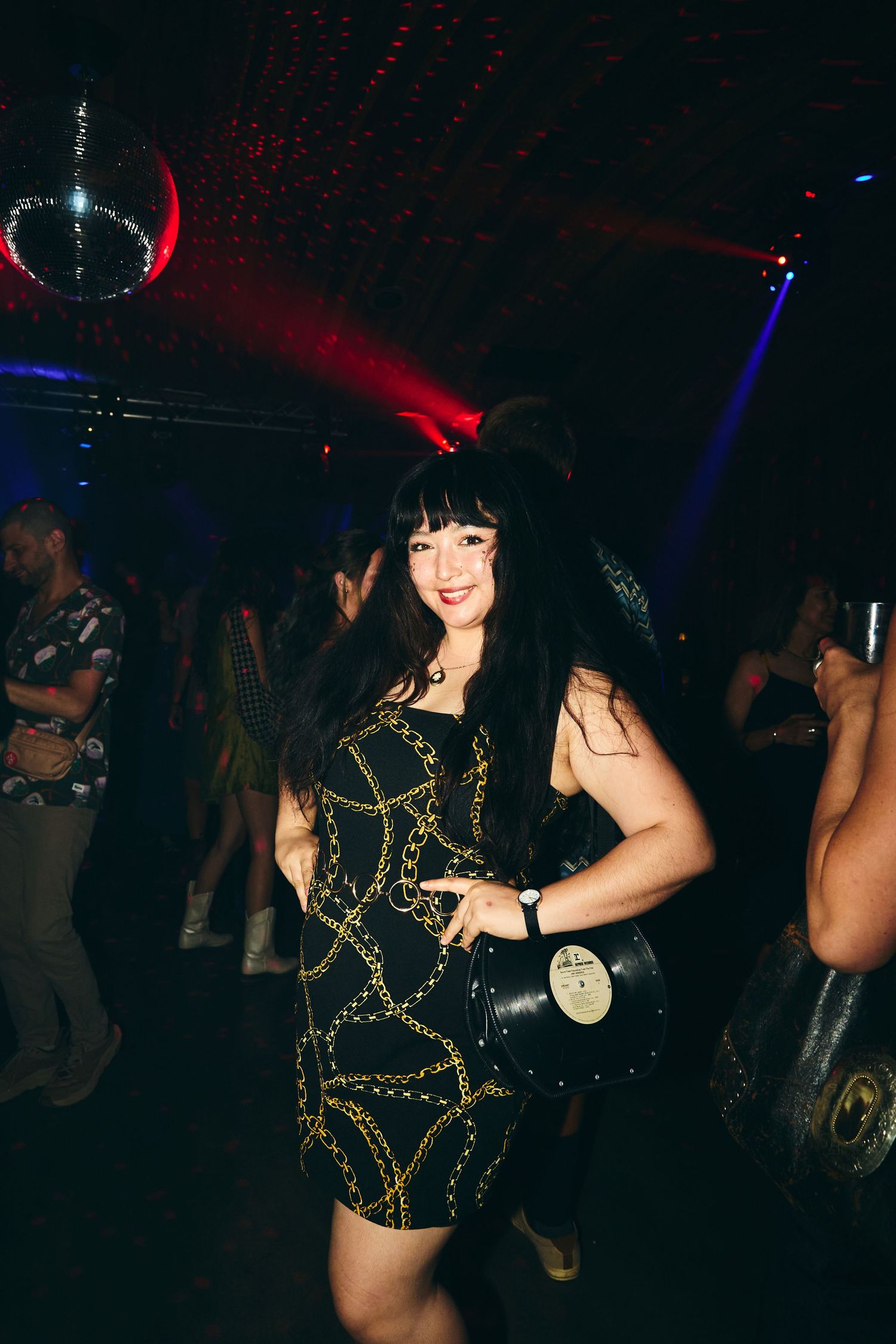 Person wearing a black and gold sequined dress dances under colorful nightclub lights and disco ball.