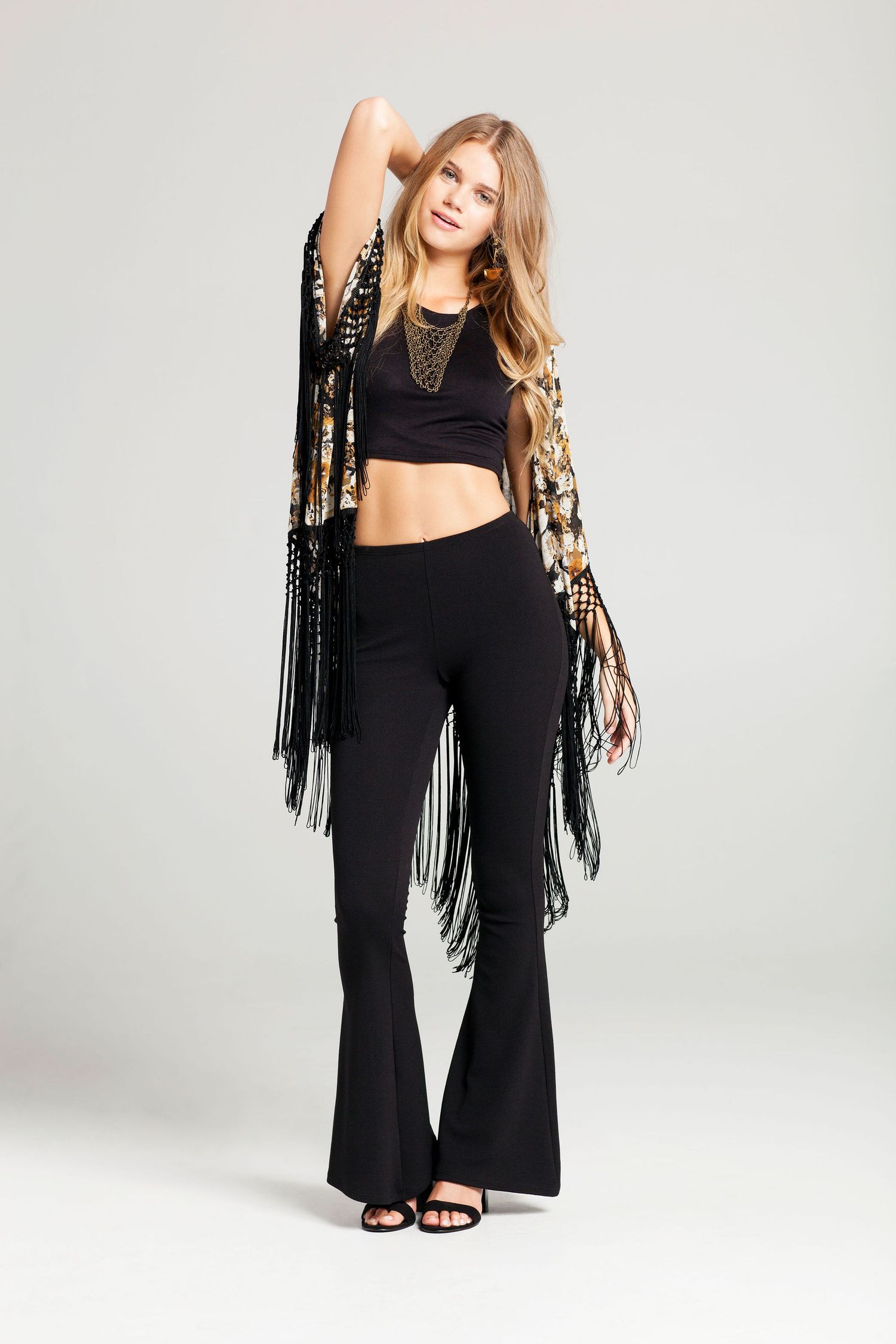 Fashion model wearing black outfit with fringed vest against grey background.