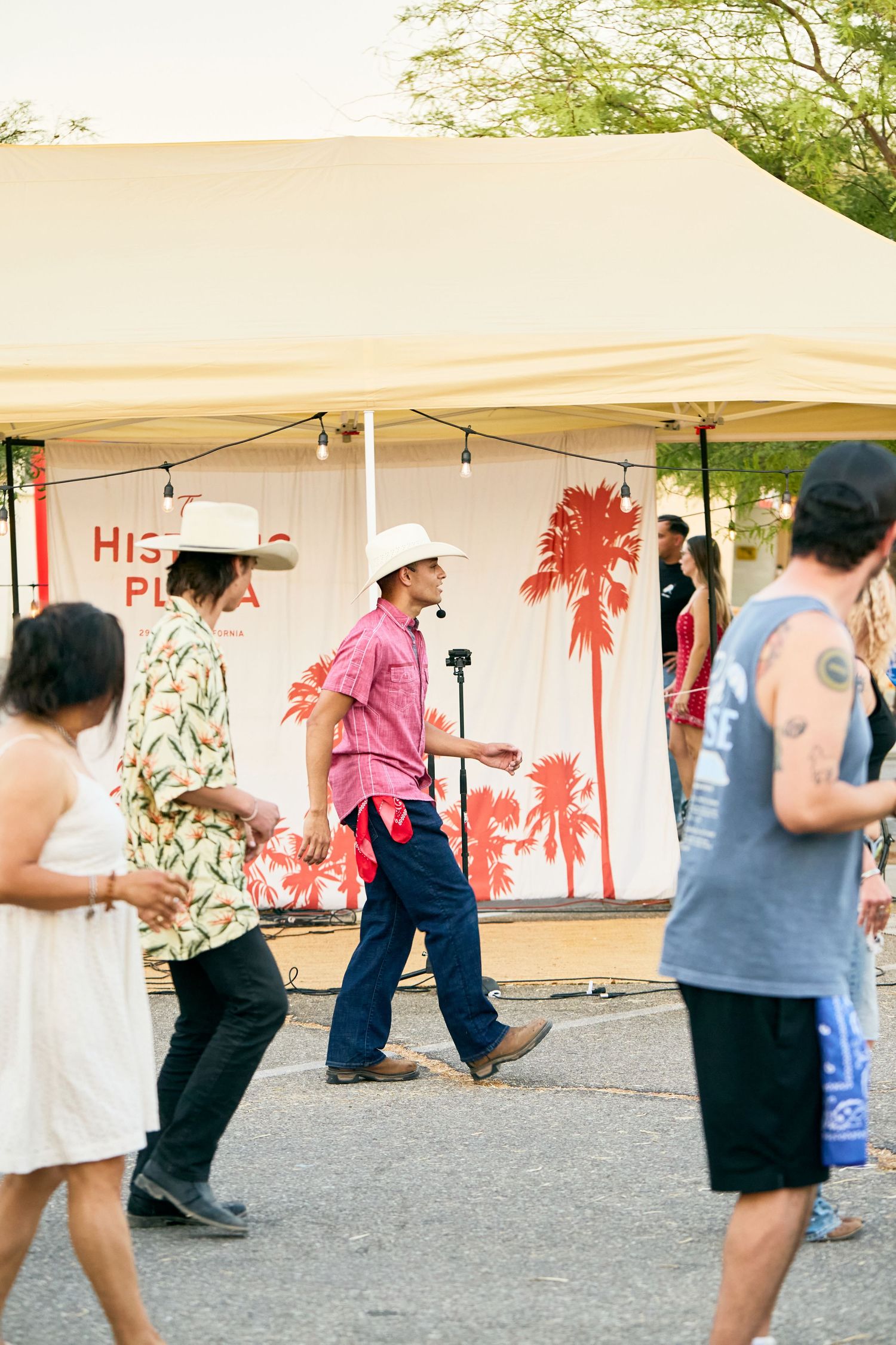 People in cowboy hats and casual attire walk near a tent with palm tree decorations at an outdoor festival.