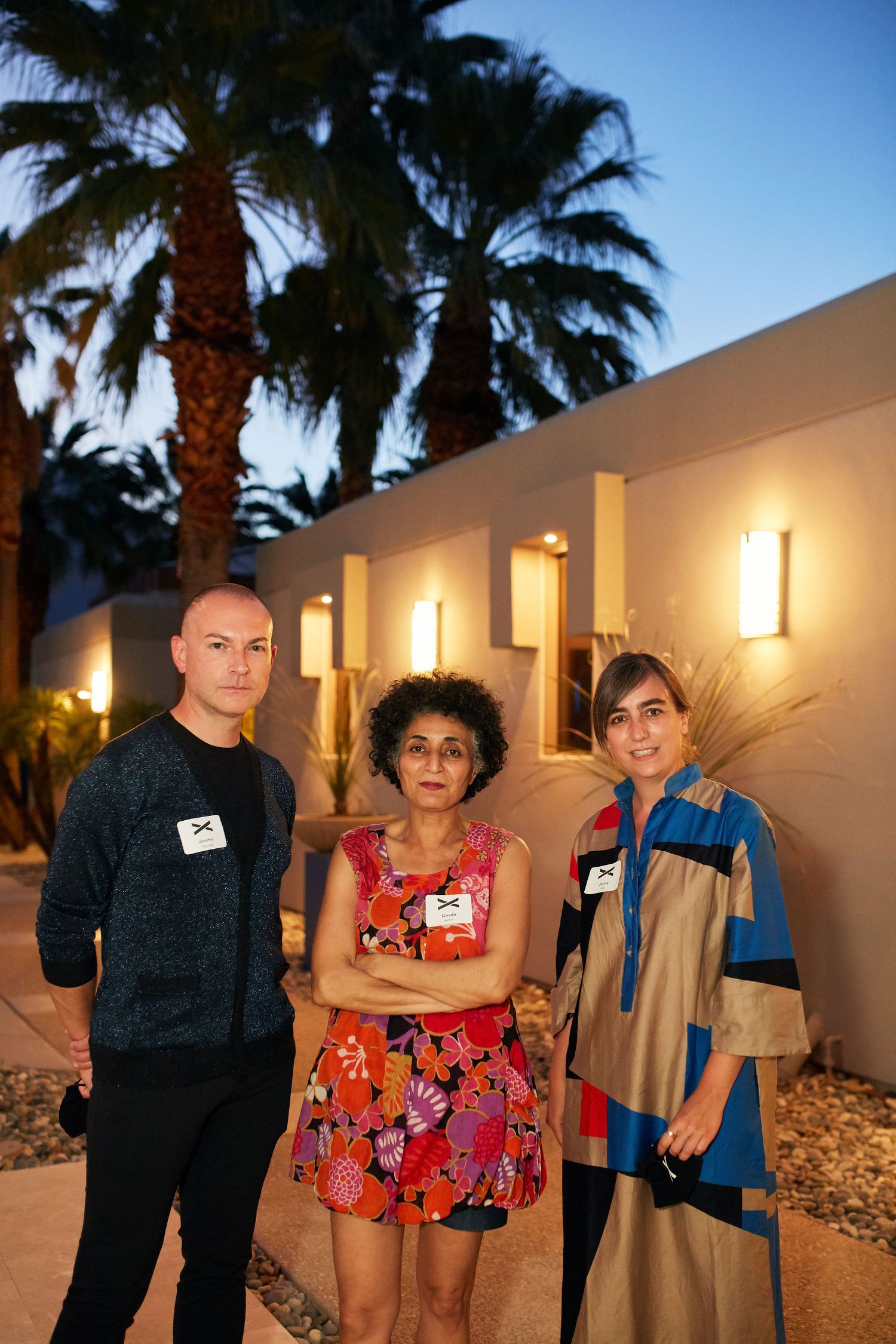 Three people stand together in front of a white building with palm trees and illuminated wall sconces at dusk.