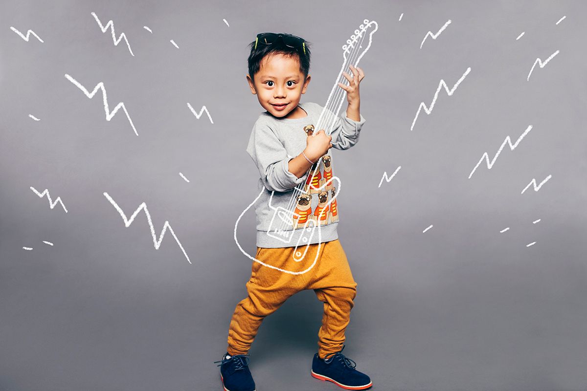 Young child wearing gray shirt and orange pants strikes a playful air guitar pose against gray background with doodle lightning bolts.