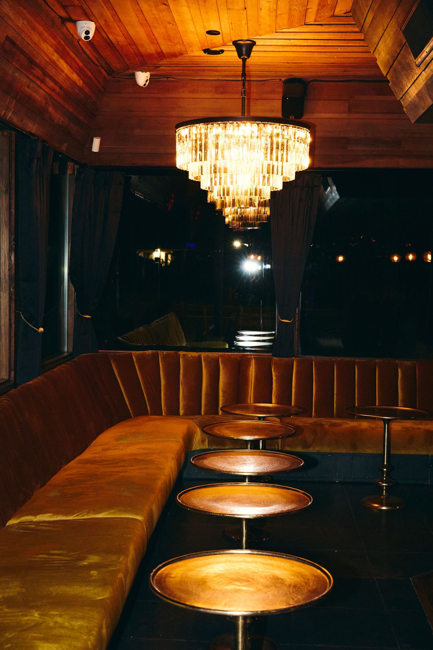 Luxurious bar interior with curved leather booth seating, round bar stools, and crystal chandelier under wood-paneled ceiling.