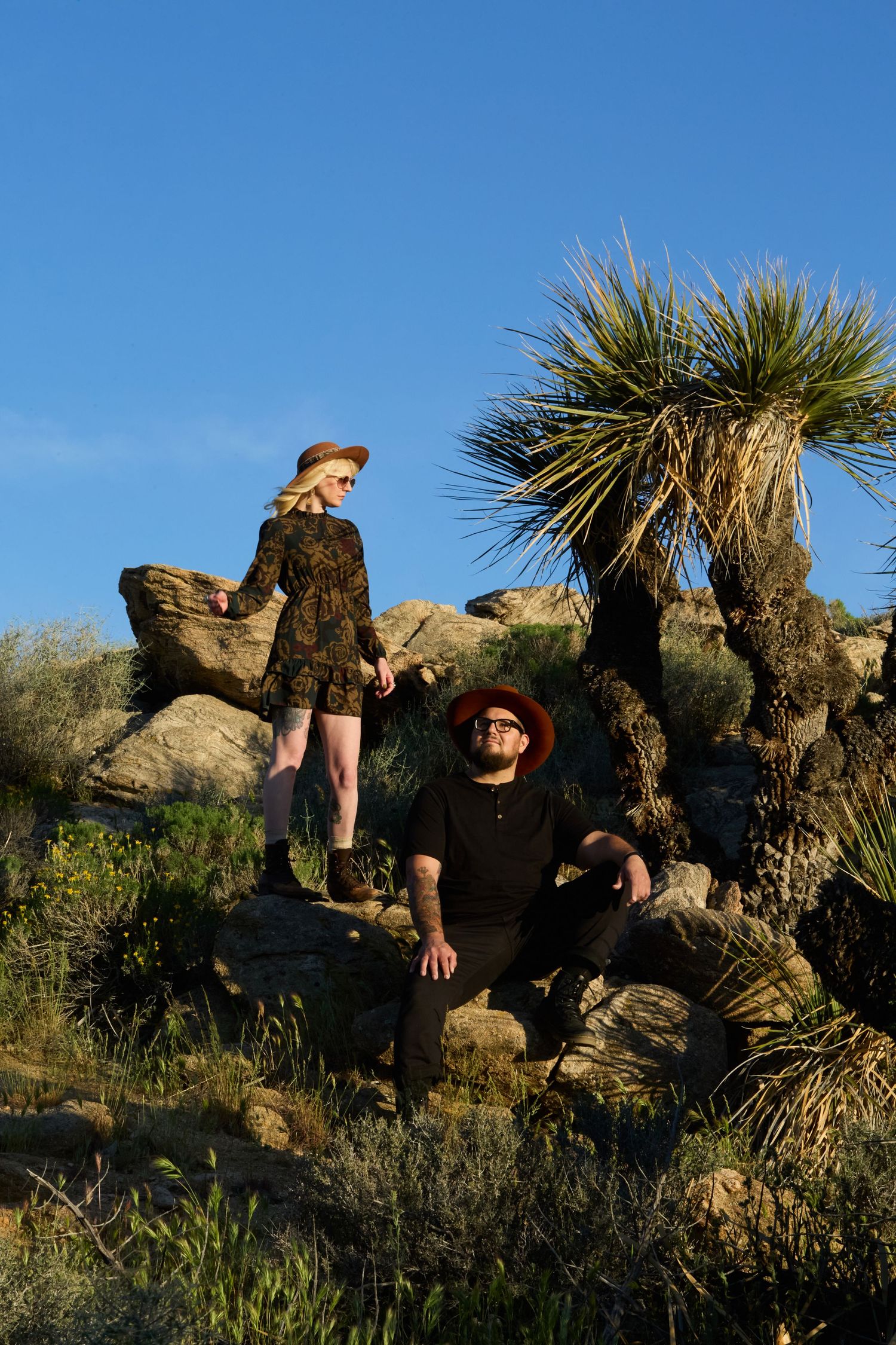 Couple posing on desert rocks near yucca plants under blue sky, woman in floral dress and hat, man in black outfit.
