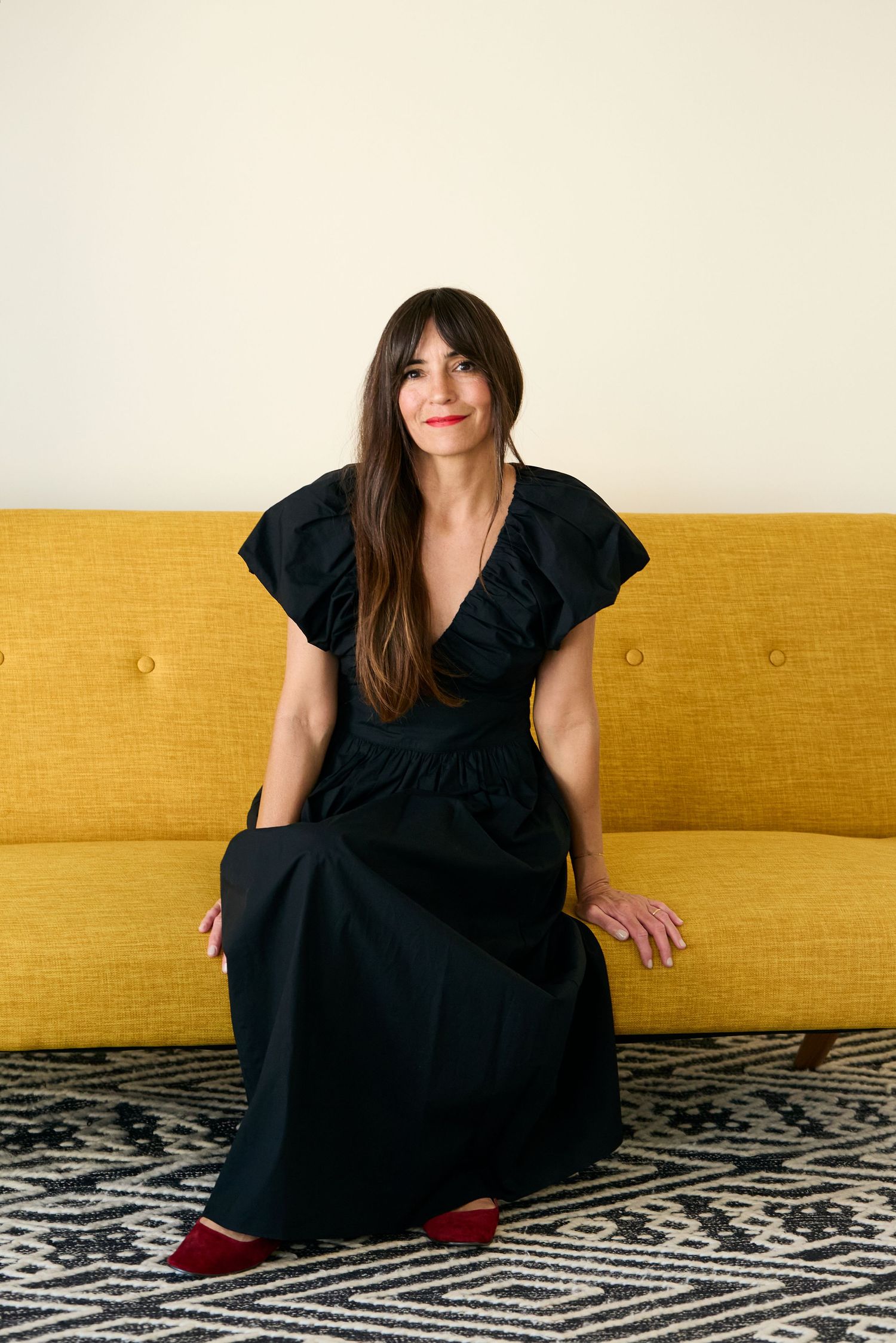 A figure in a black dress sits on a yellow couch against a geometric patterned rug in a lifestyle photo series.