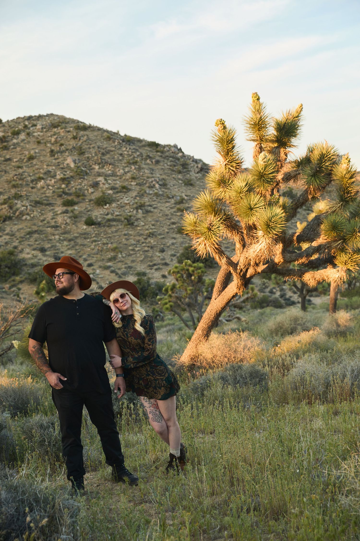 Couple in brown hats standing in desert landscape with Joshua tree and hills at golden hour.