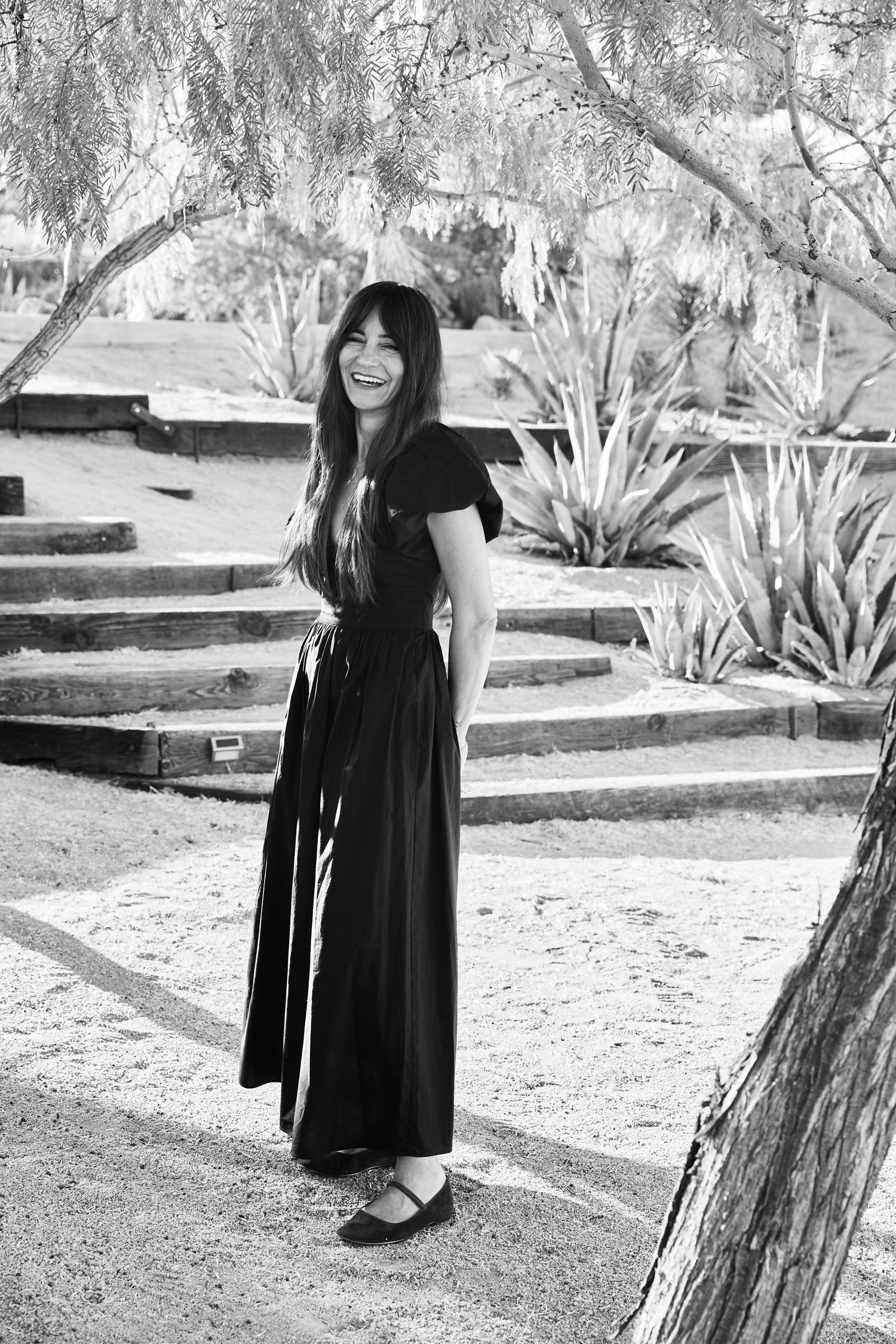 Black and white portrait of someone in a flowing black maxi dress standing on garden steps surrounded by agave plants.