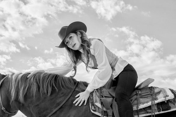 Black and white photo of a cowgirl leaning over a saddled horse against a cloudy sky.