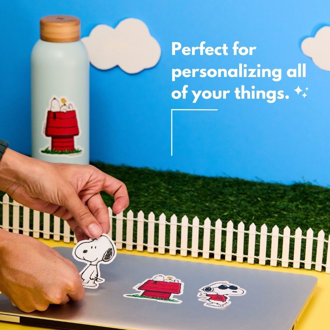 Hands placing Snoopy cartoon stickers on a decorative scene with white picket fence and blue sky.