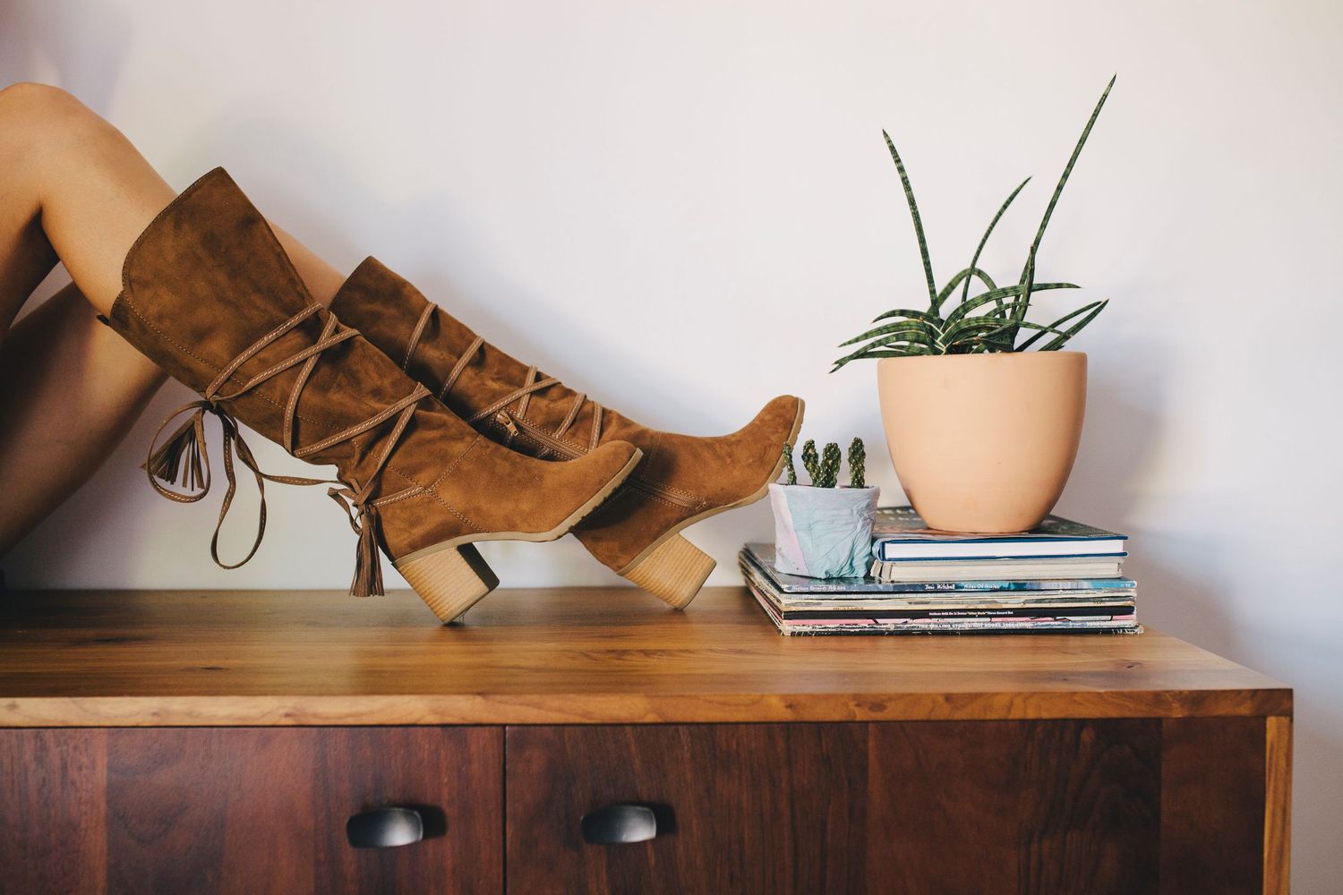 Tall brown leather boots displayed on wooden dresser next to potted plant and stacked books.