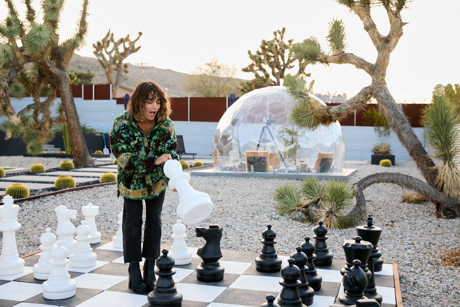 A collection of outdoor chess pieces and tables arranged in a desert landscape with Joshua trees and mountains in the background.