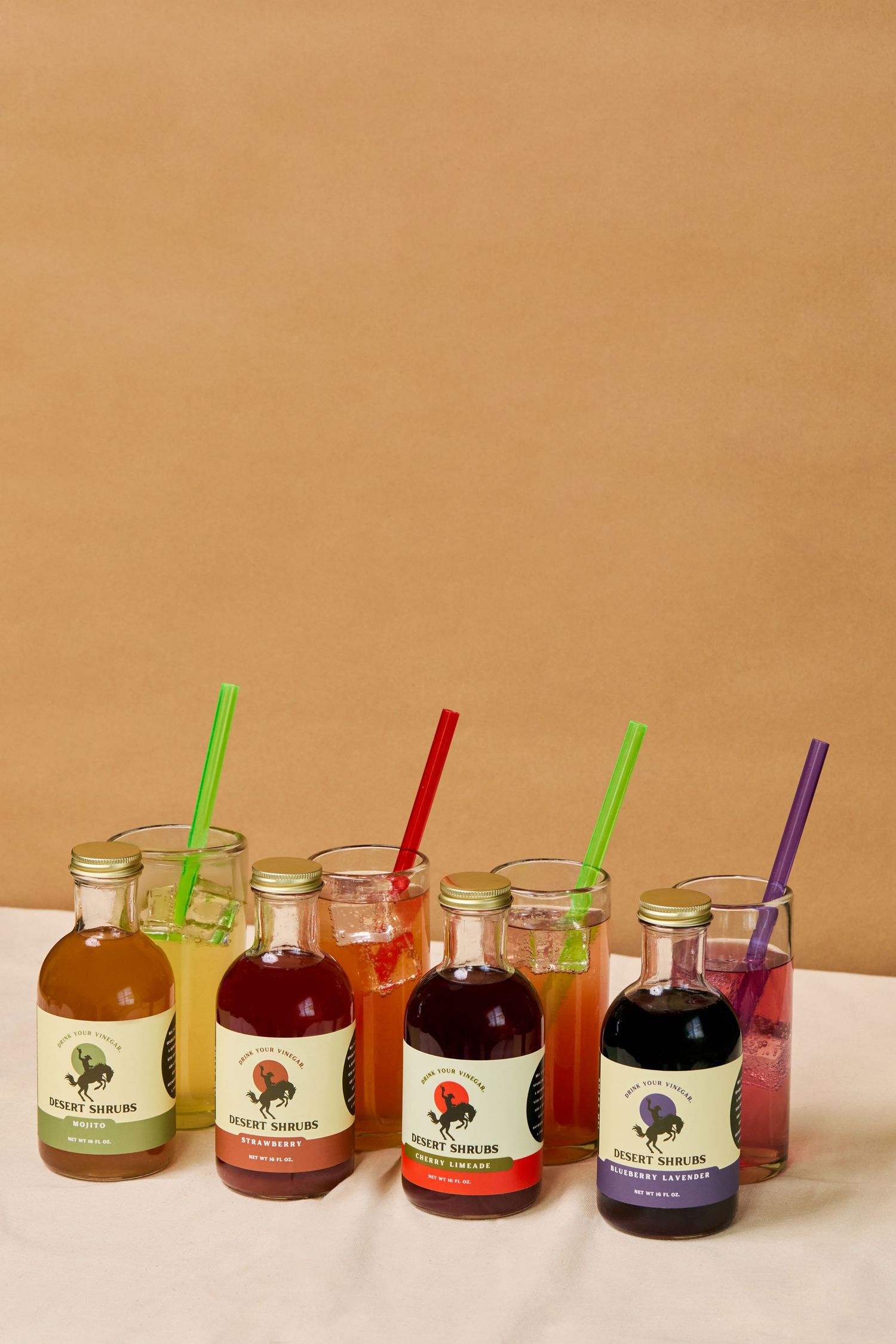 Four Desert Suburb craft syrup bottles with colorful drinks in glasses with straws on a white cloth.