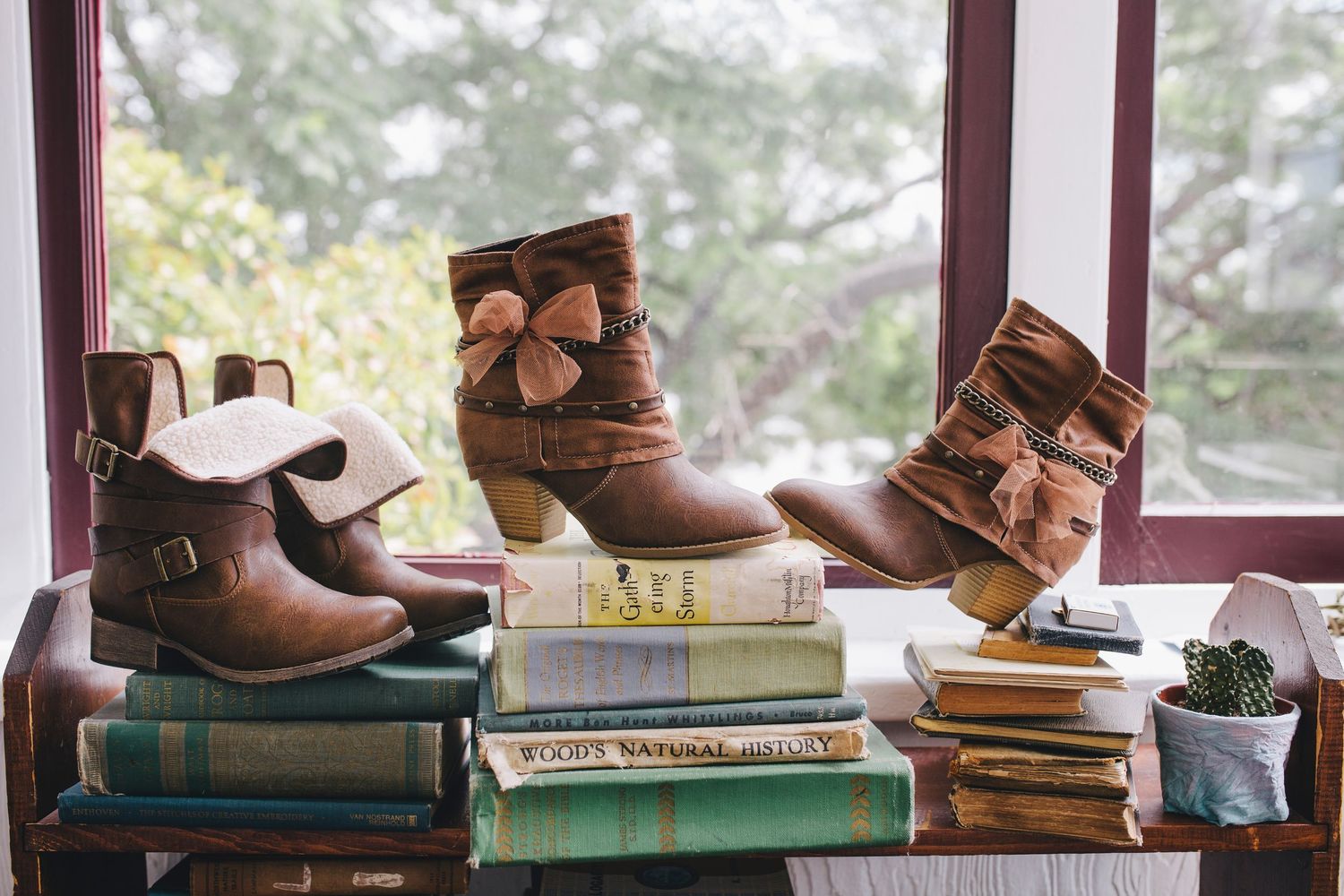 Multiple pairs of brown leather shoes and boots arranged in a line on stacked books near a window sill.