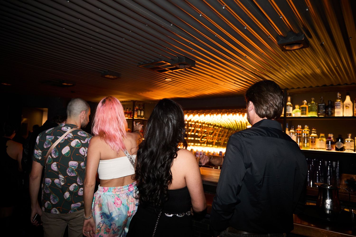 Group of people socializing at a dimly lit bar with warm ambient lighting and wooden slat ceiling.