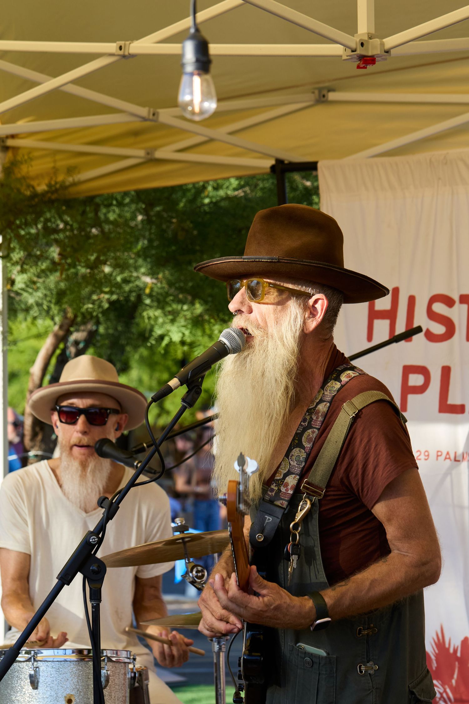 Elderly bearded musician in brown hat plays guitar and sings at outdoor festival stage with drummer performing behind him.