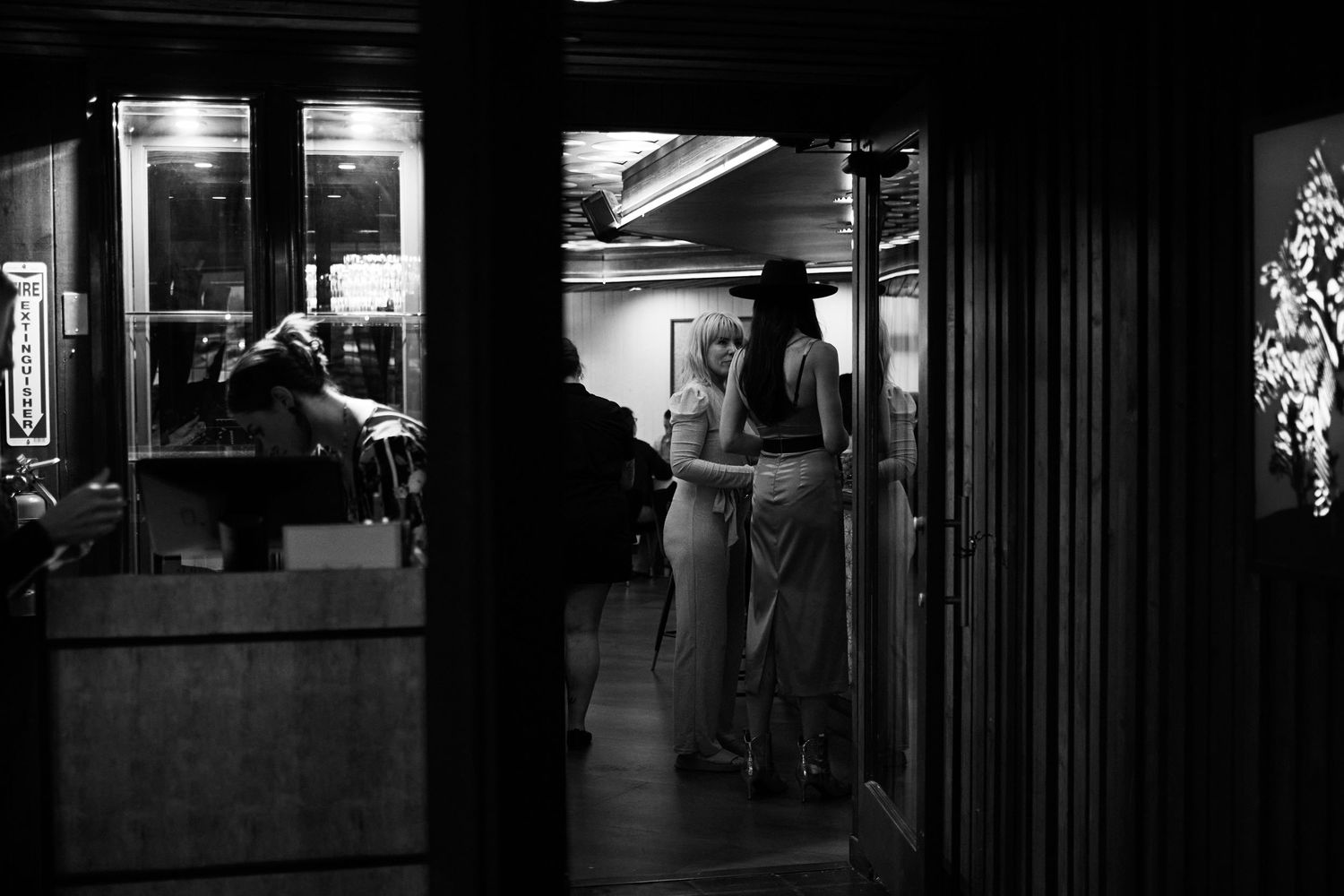 A moody black and white nighttime scene through a restaurant window shows illuminated silhouettes of people inside.