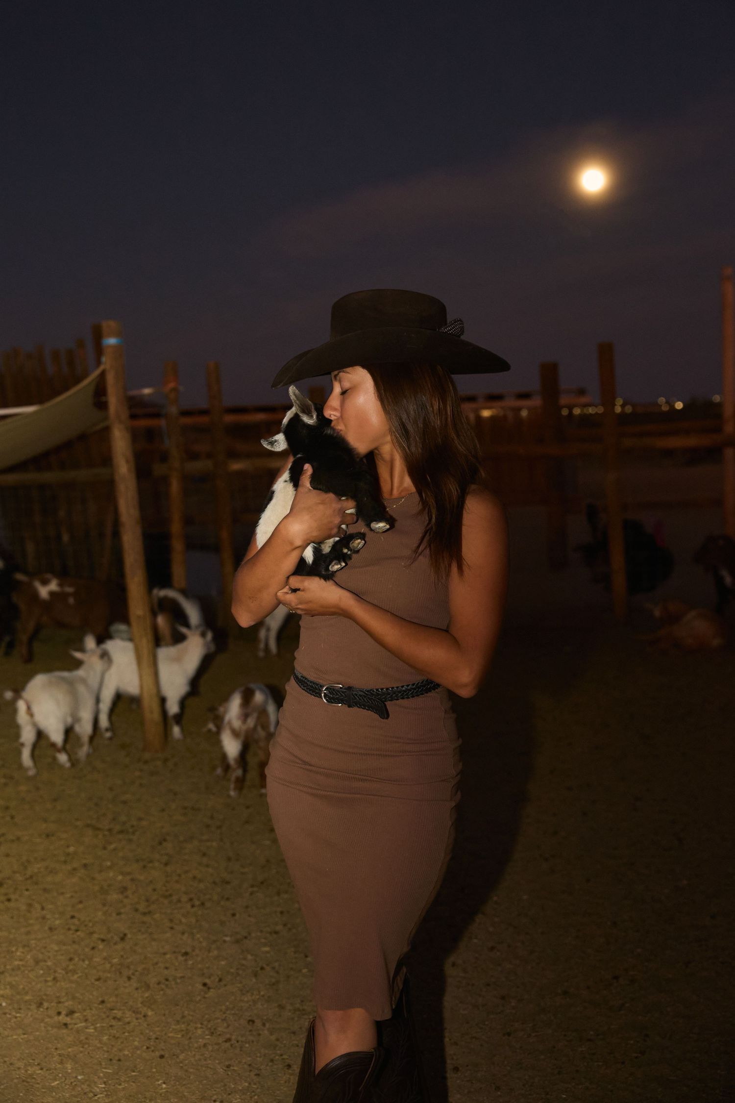 Woman in brown dress and hat kisses baby goat at night farm under full moon.