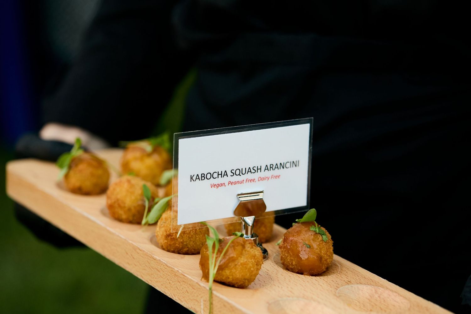 A wooden serving board displays crispy golden croquettes with a small sign holder for menu descriptions.