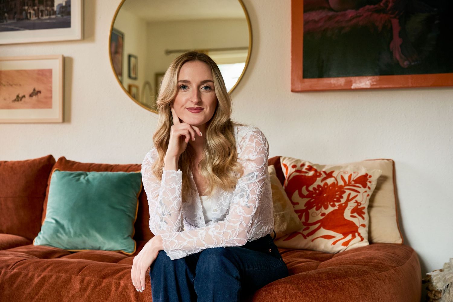 Blonde woman in white lace top sits on rust velvet sofa with colorful pillows, posing thoughtfully in stylish living room.