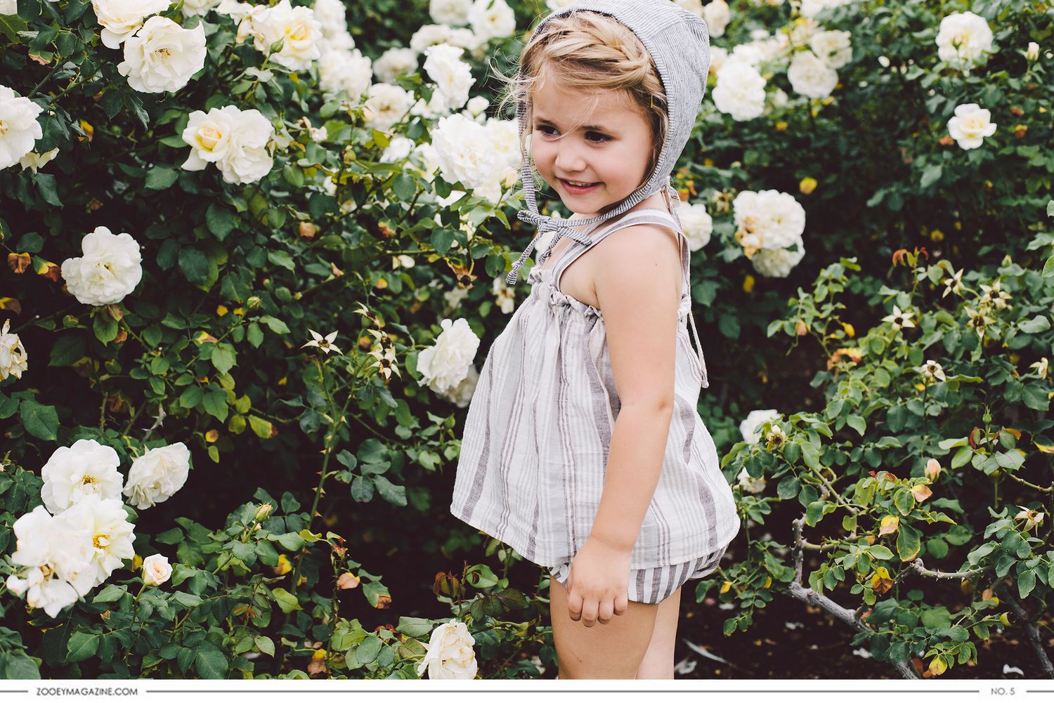 A young child in a summer dress stands in front of a blooming white rose bush garden.