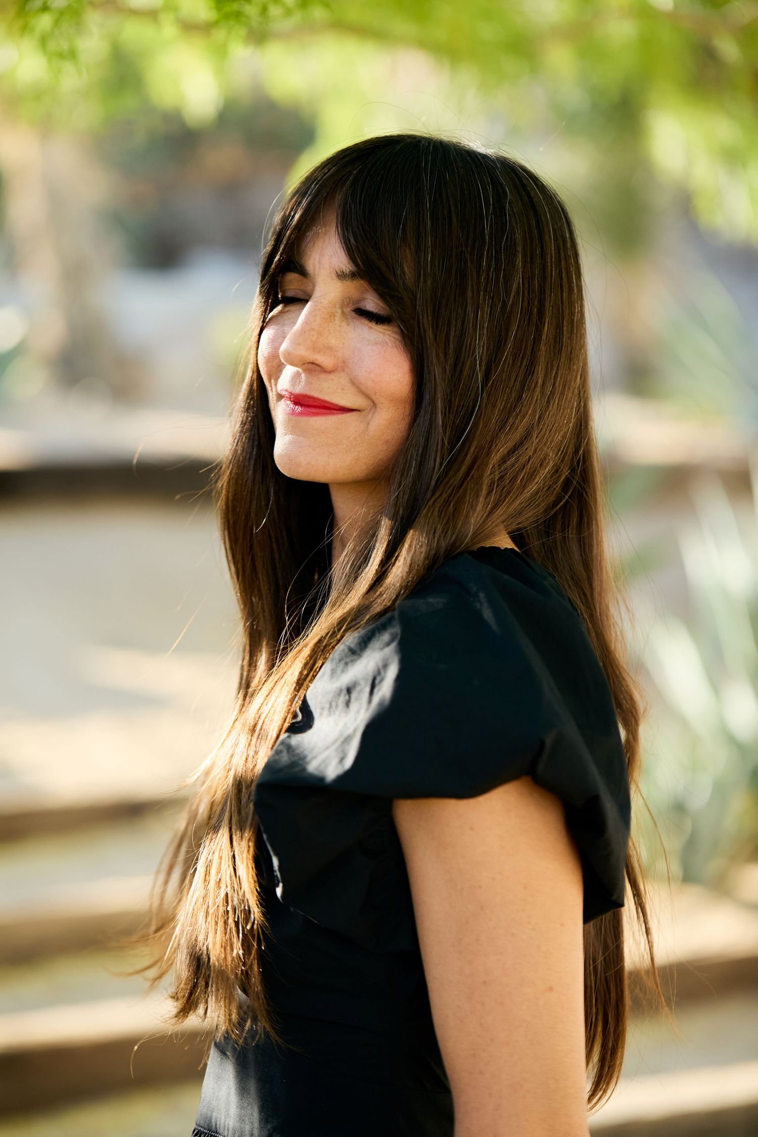 Woman with long dark hair and red lipstick smiles in outdoor setting while wearing black top.