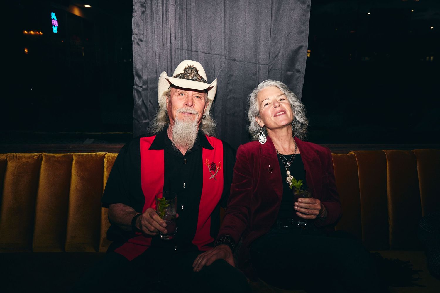 Two people in red attire sit together on a brown velvet bench holding drinks at what appears to be a bar or lounge.