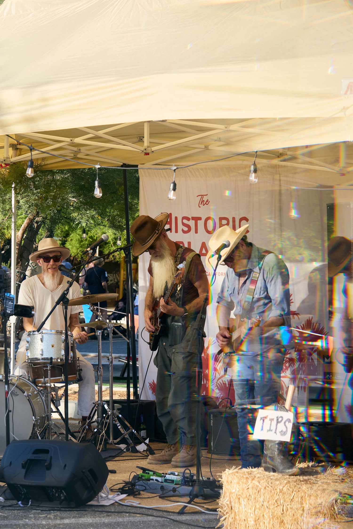 Live band performs under tent at Historic Plaza outdoor festival, featuring guitarist, drummer, and tips jar on hay bale.