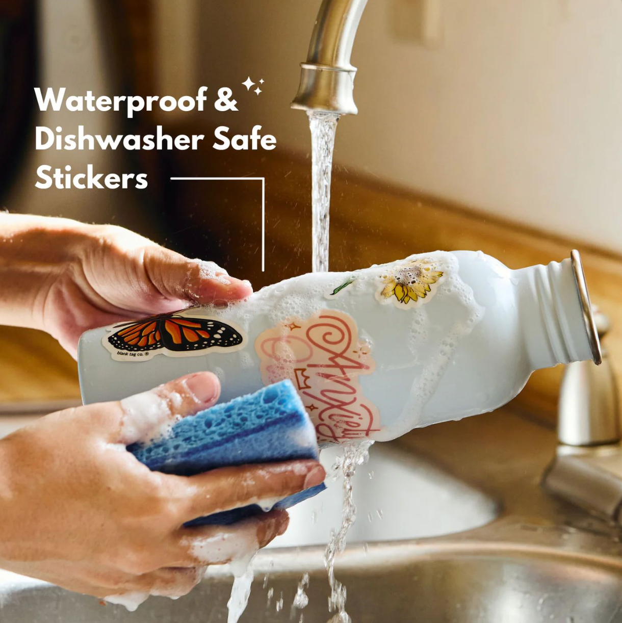 Person washing a white water bottle decorated with butterfly stickers using a blue sponge under running water.