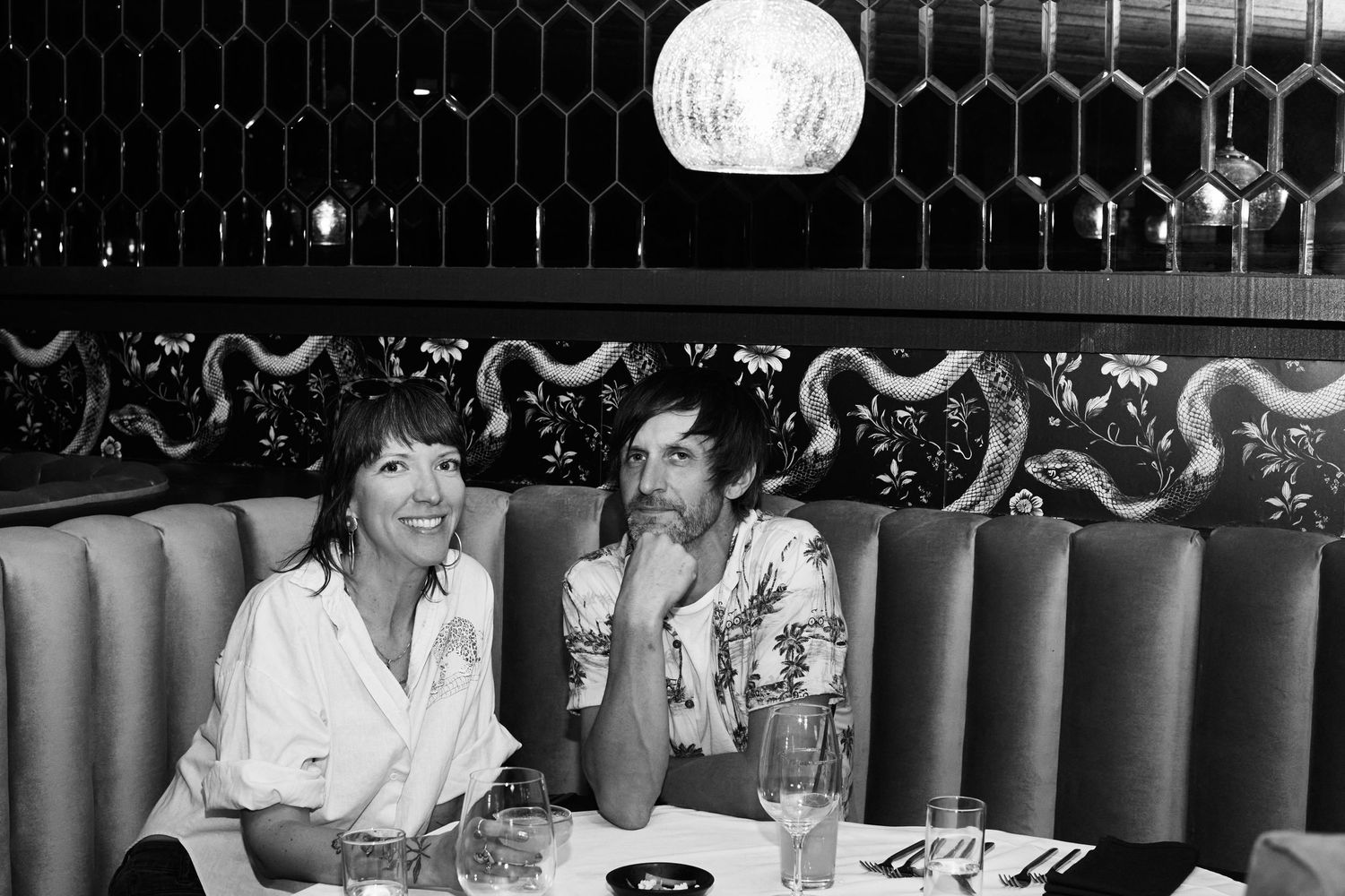 Two people sit at a booth in a retro nightclub with a patterned wall, having drinks and engaging in conversation.