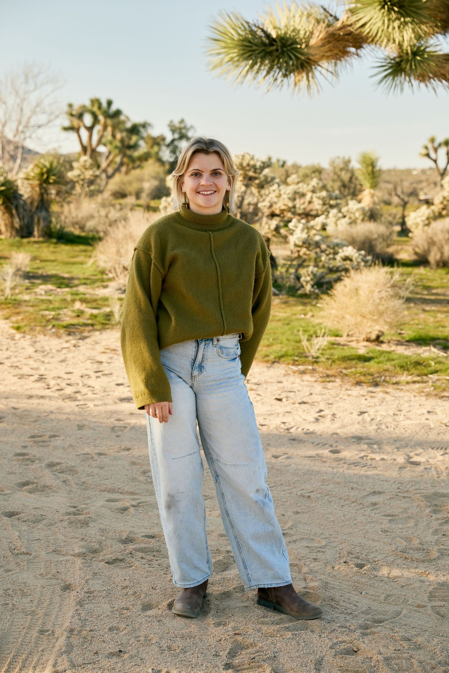 Person wearing olive green sweater and light wash jeans poses outdoors in desert landscape with Joshua trees.