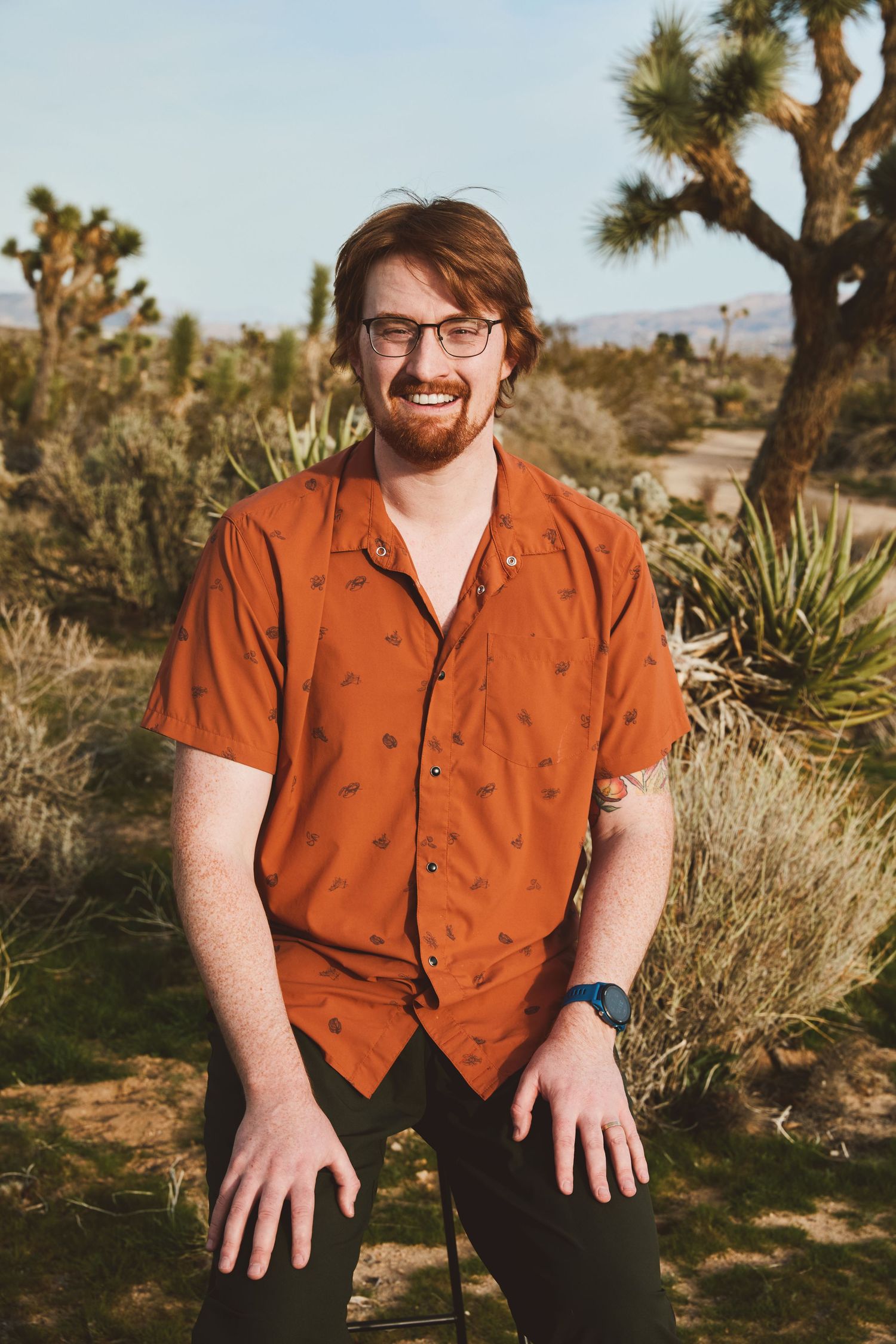 Person wearing an orange button-up shirt poses outdoors in a desert landscape with Joshua trees in the background.