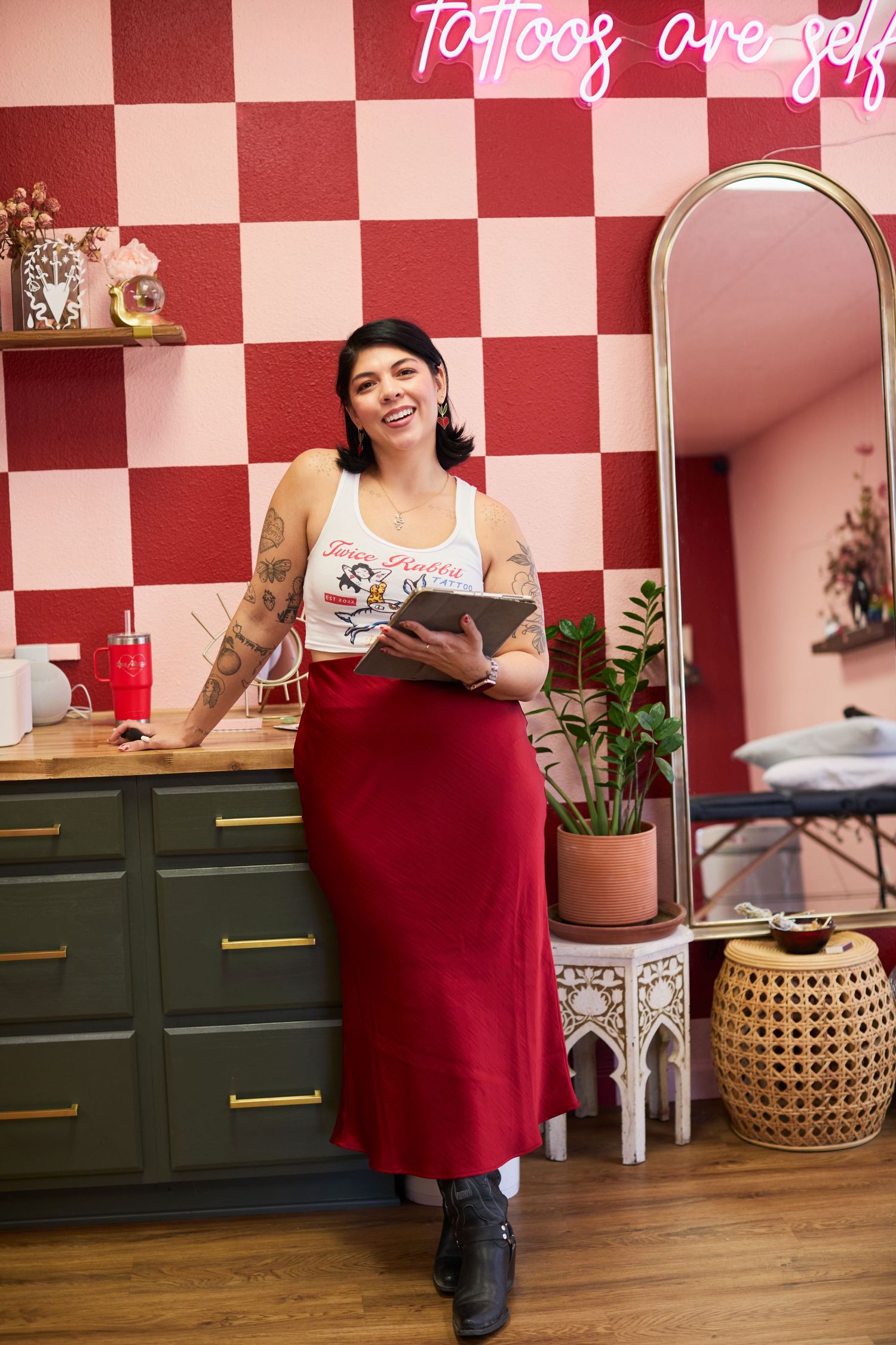 Smiling tattooed woman in red skirt holds tablet in a stylish tattoo studio with checkered walls and neon sign.