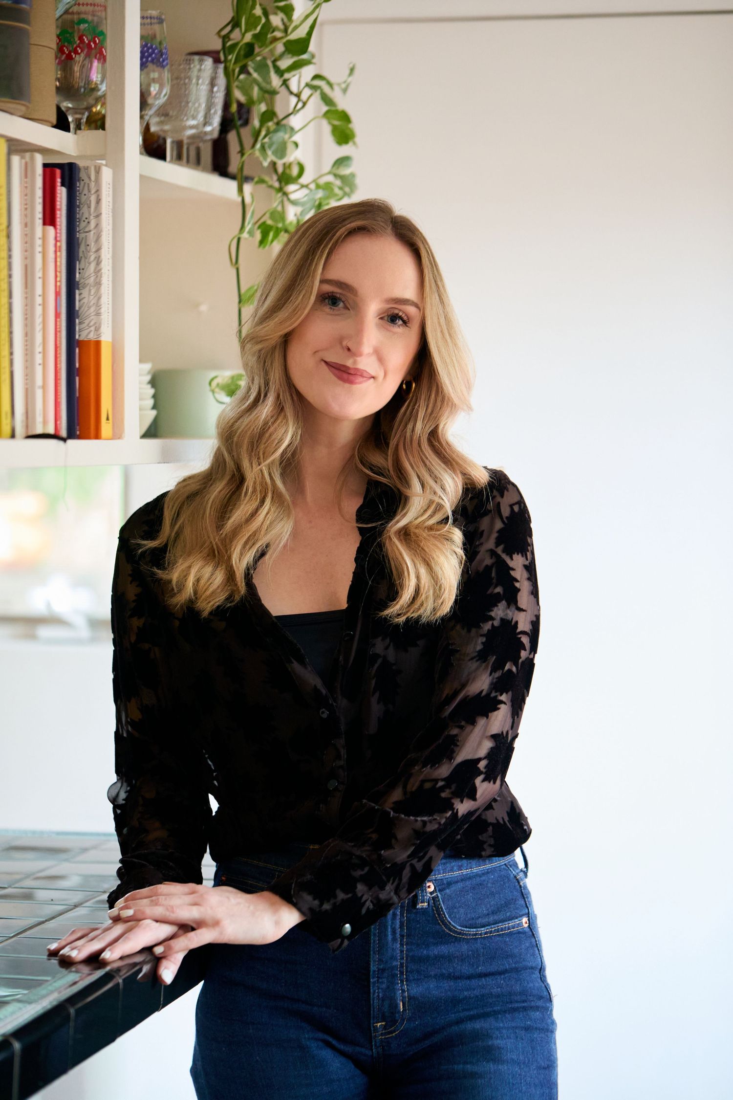 Smiling blonde woman in black lace jacket and jeans poses in front of white bookshelf with plants.