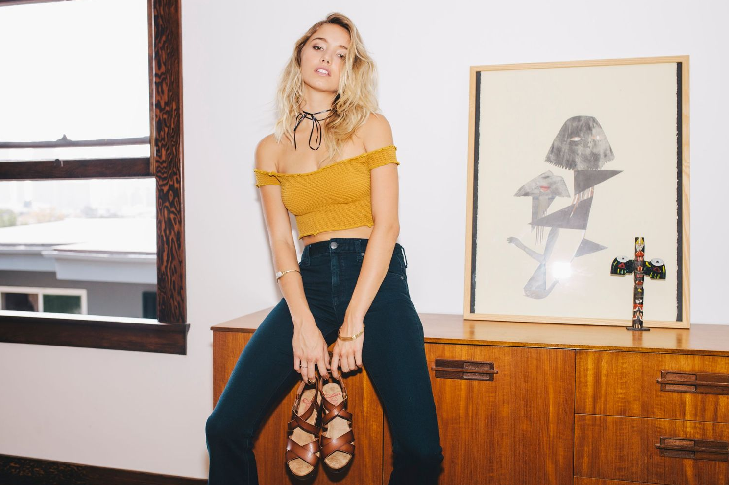 Yellow top and dark pants outfit styled with sandals against white wall with framed artwork.
