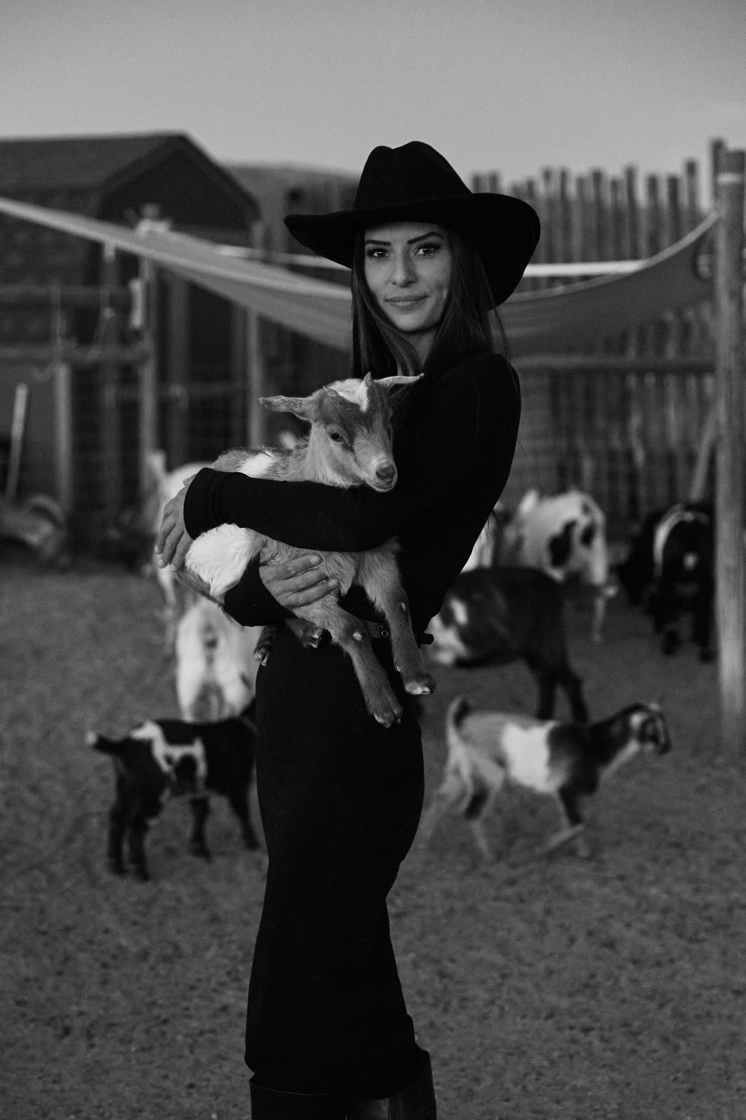 Woman in black cowboy hat holding a baby goat surrounded by goats at a farm in black and white.