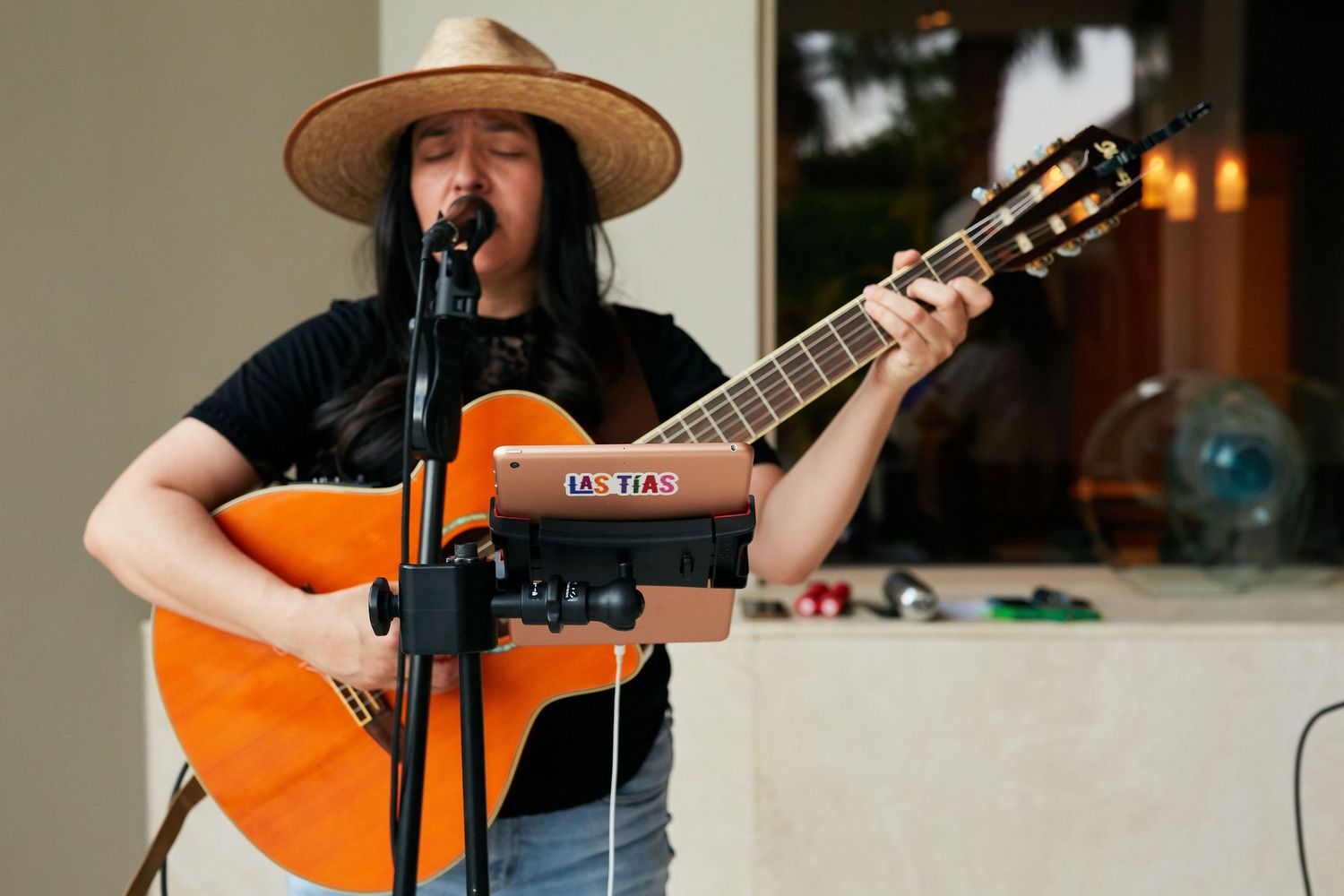 A musician performs with an acoustic guitar outdoors while wearing a wide-brimmed hat and standing at a microphone.