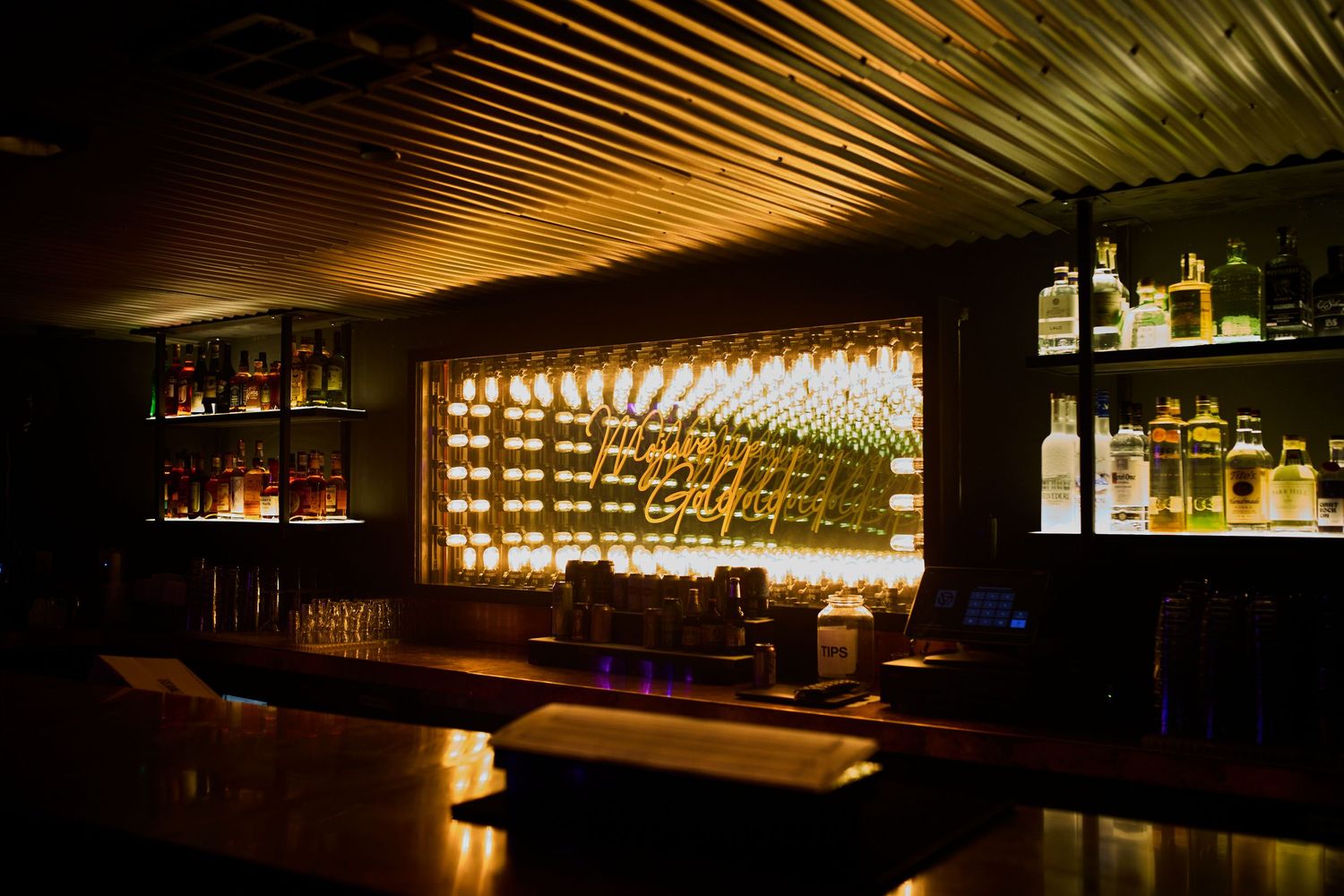 Interior of a dimly lit upscale bar with illuminated backlit shelving displaying bottles and modern architectural lighting design.