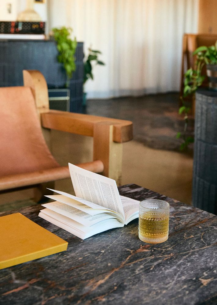 A glass of whiskey sits beside an open book on a dark marble table with plants in the background.