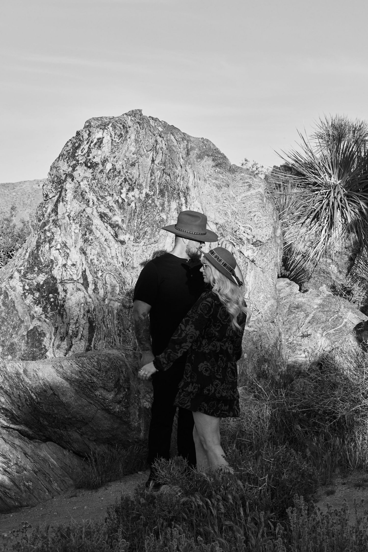 Black and white photo of a couple in hats embracing near rocky desert landscape with Joshua trees.