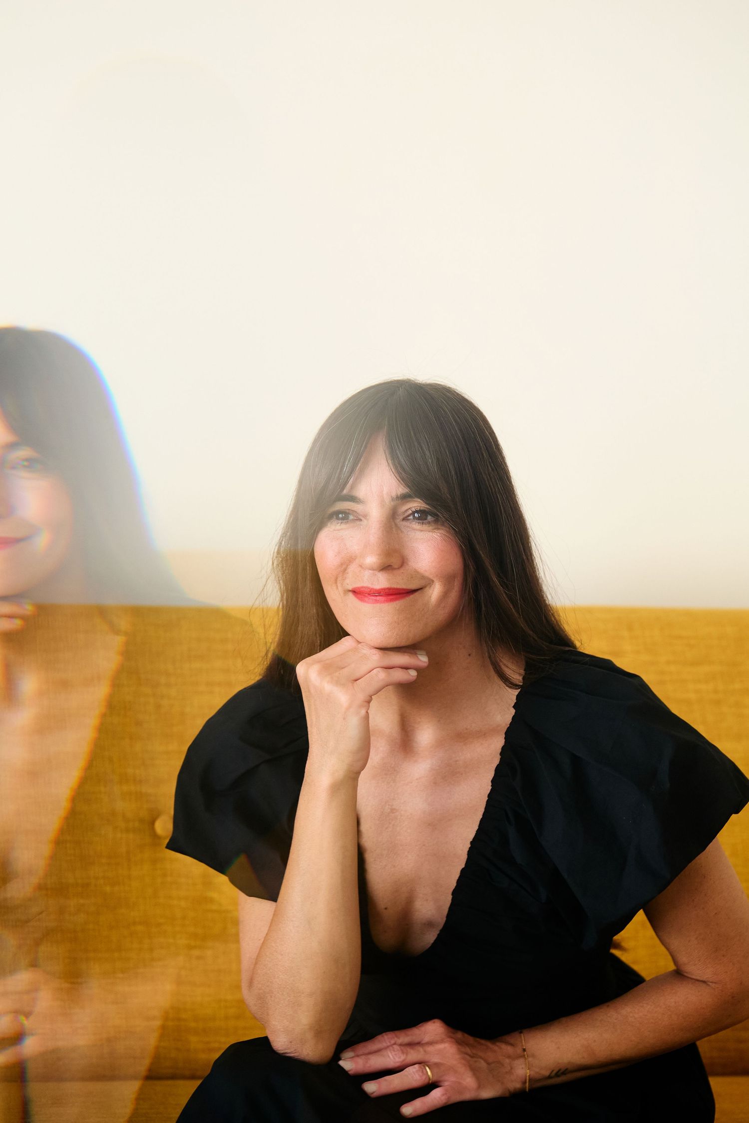 Cheerful person in black dress sits on yellow sofa with reflection creating artistic double image effect.