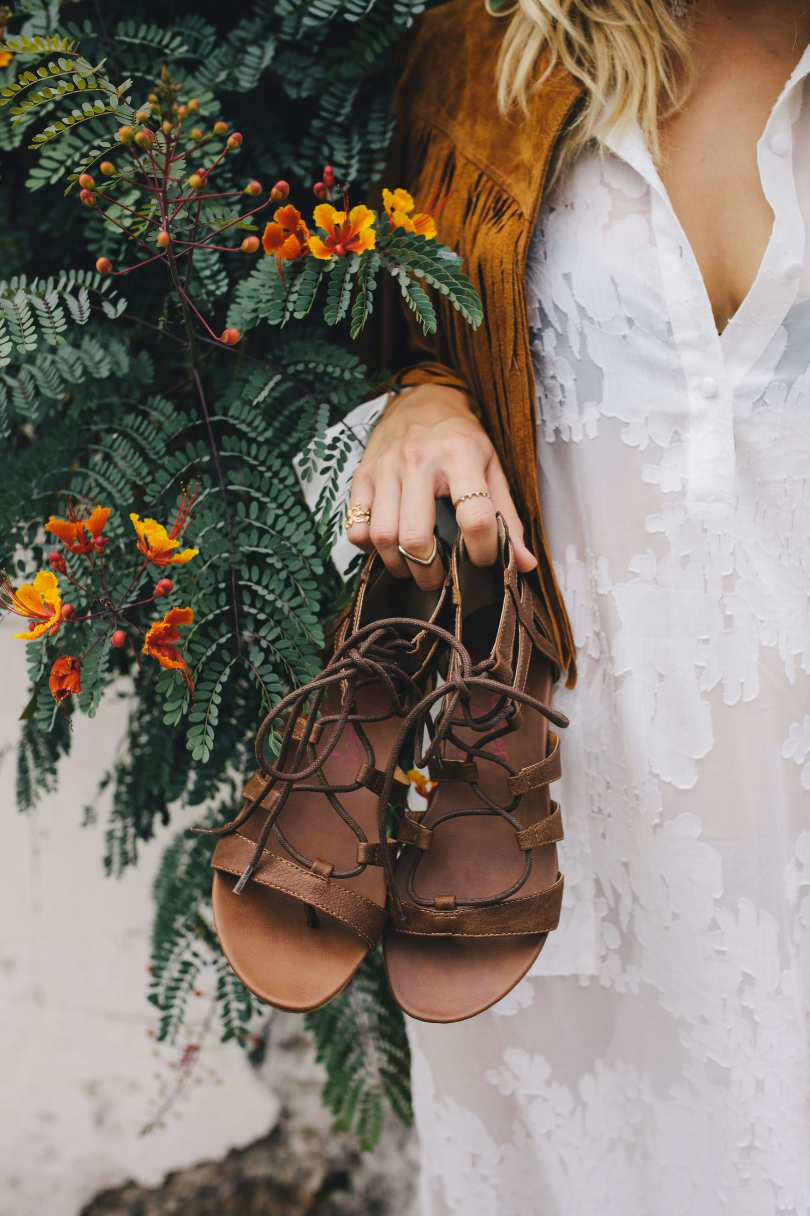 Brown leather lace-up sandals held against white dress and flowering plant background.